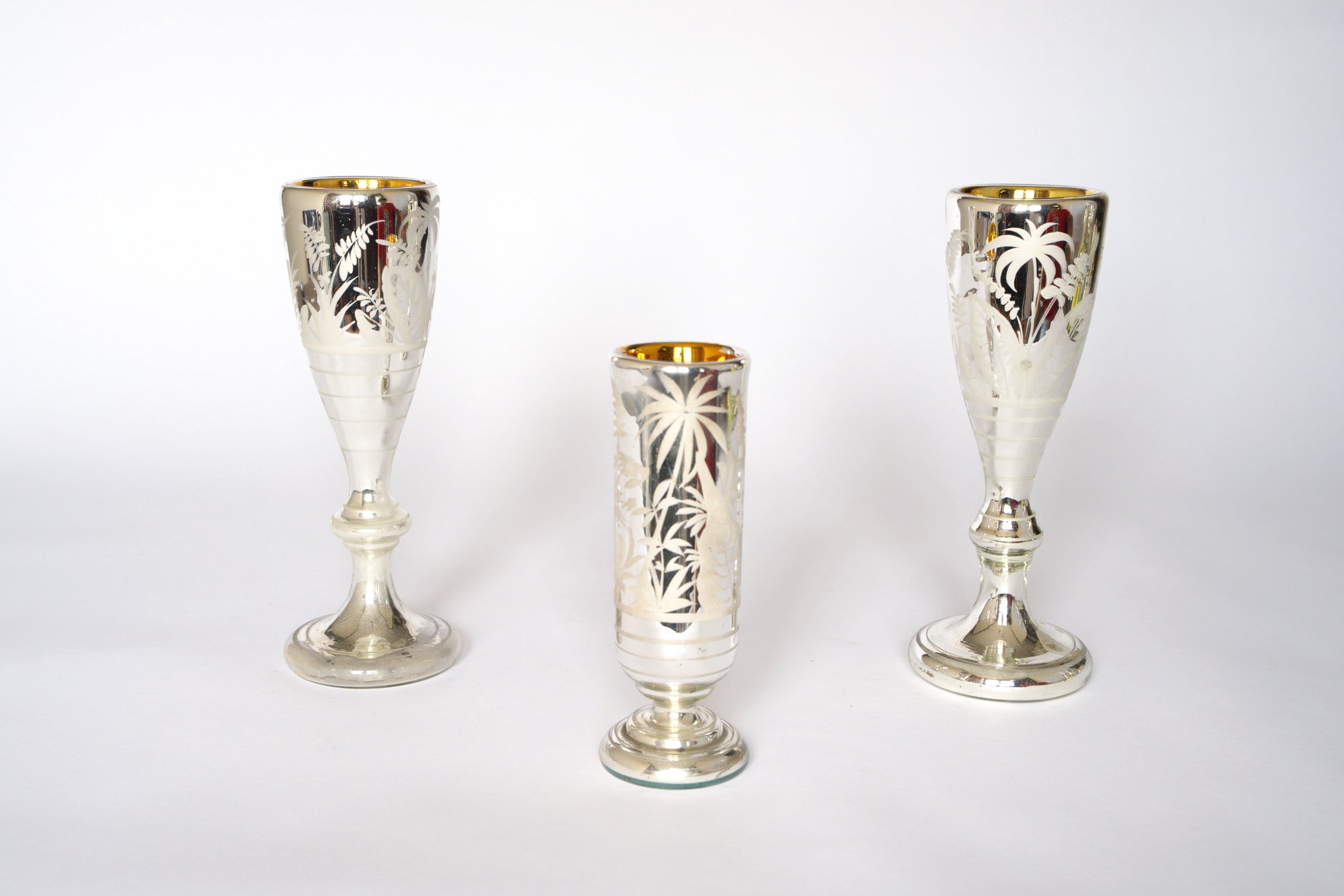 The Vintage Etched Mercury Glass Vessel Set by Euro Trash features three ornate silvered vessels with engraved palm tree motifs on a white background. The central piece is shorter and cylindrical, flanked by two taller, wider-based vessels.