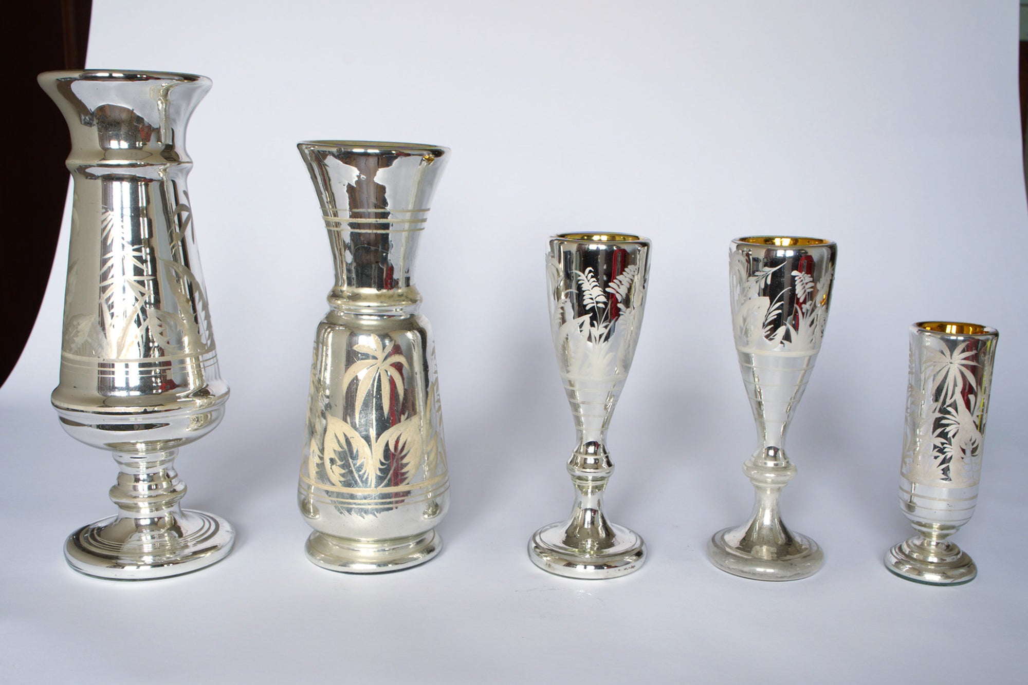 The Euro Trash Vintage Etched Mercury Glass Vessel Set features five silver vases and goblets of varying heights, each with botanical motifs and a reflective finish, arranged in a row on a white background.