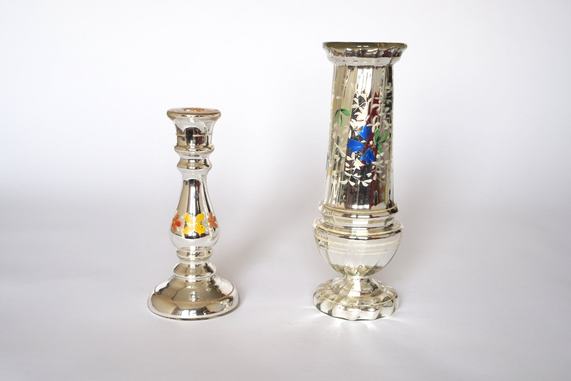 The Pair of Vintage Silver Mercury Glass Vases by Euro Trash, one with orange floral details and the taller with blue and white flowers, adds striking sculptural décor against a plain white backdrop.
