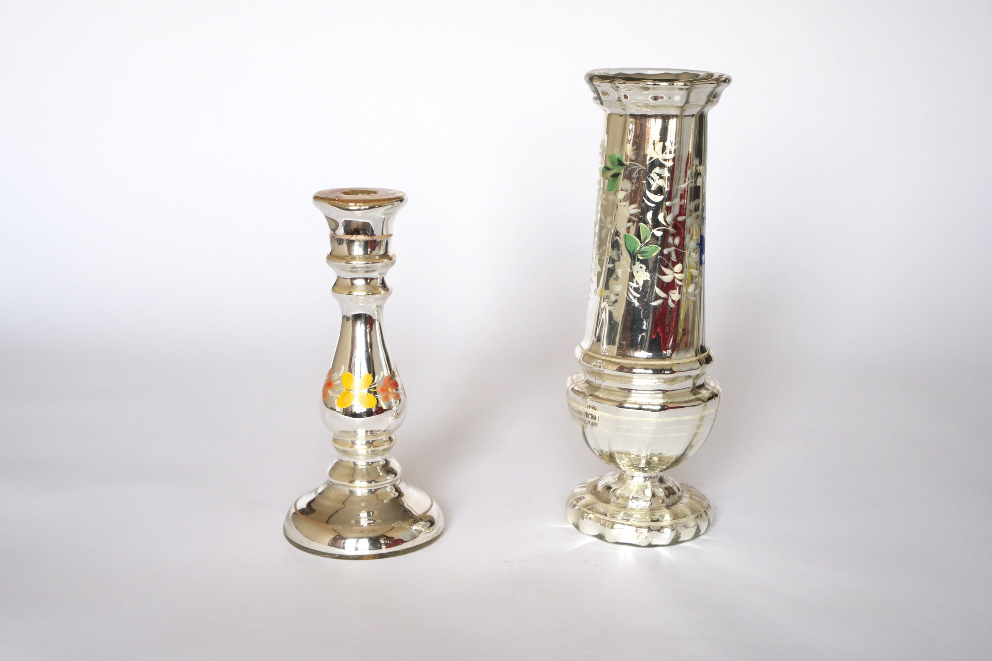 Two vintage silver mercury glass vases by Euro Trash on a white background: one features yellow and orange floral accents, the other is taller with ornate, hand-painted floral detailing.