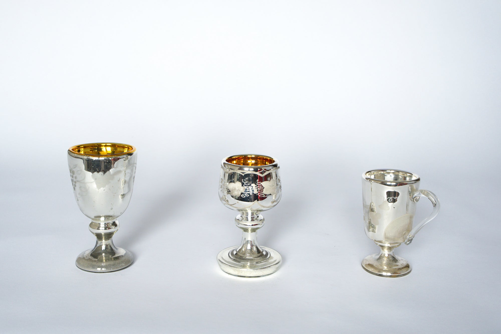 The Euro Trash Vintage Mercury Glass Stemmed Cup Set features three silver cups with gold interiors in distinct shapes and bases, including one with a handle, all displayed side by side on a plain white background.