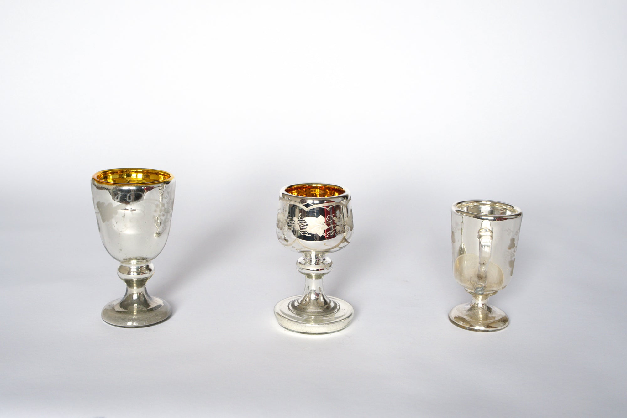 The Euro Trash Vintage Mercury Glass Stemmed Cup Set features three uniquely shaped cups with gold interiors and etched details, displayed side by side on a white background—an ideal decorative accent for any collection.