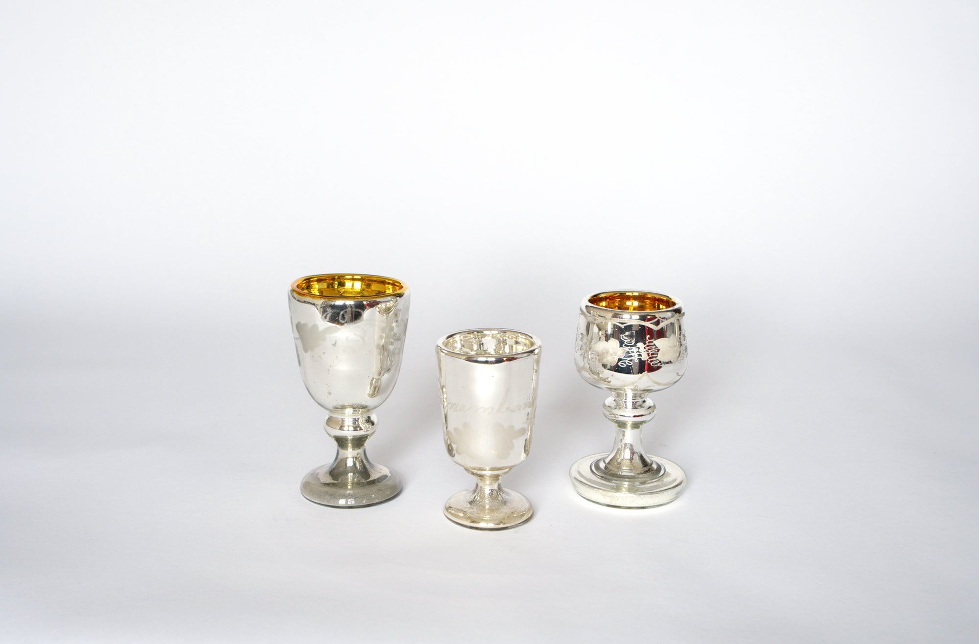 The Euro Trash Vintage Mercury Glass Stemmed Cup Set features three etched cups of varying heights and shapes, each with a gold interior and subtle tarnish. Displayed side by side on white, they're a charming decorative accent.