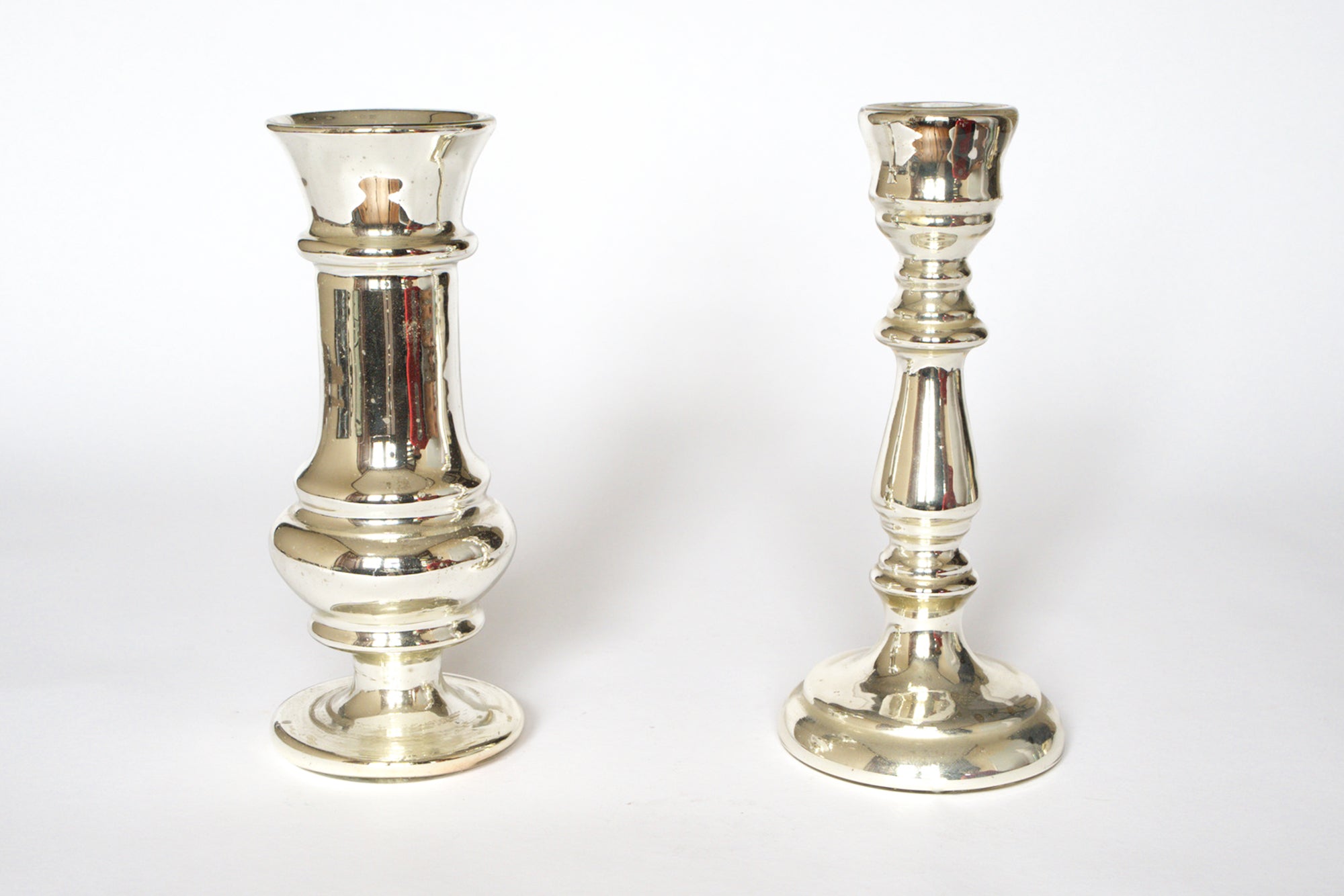 A pair of Vintage Mercury Glass Candleholders by Euro Trash stands upright on a white background. The left candleholder is taller with a wider base, while the right one is shorter and slimmer. Both feature a reflective silvered finish.
