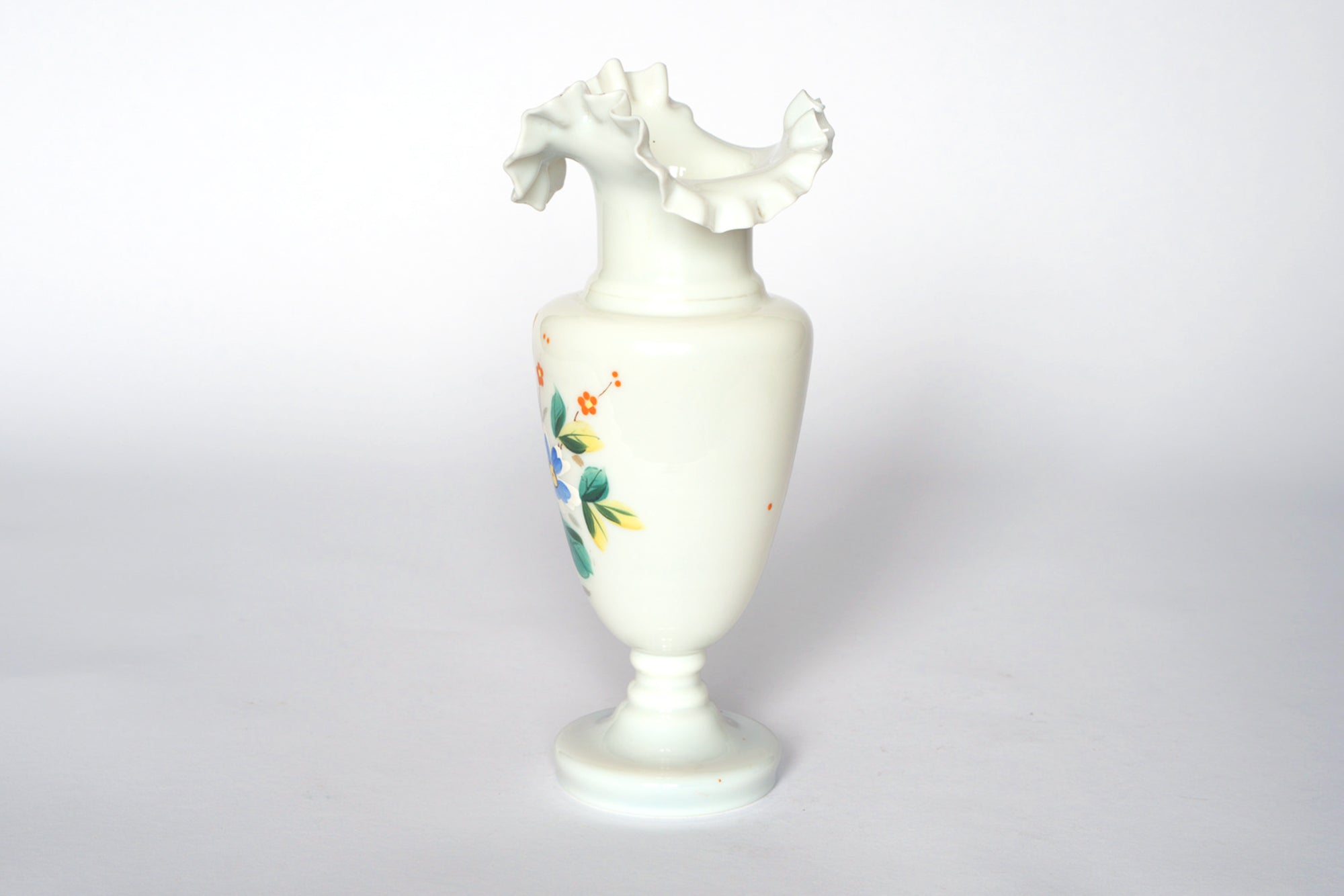 The Vintage Hand-Painted Milk Glass Vase by Euro Trash features a ruffled, flared rim, hand-painted floral design, and a round pedestal base, shown against a plain white background.