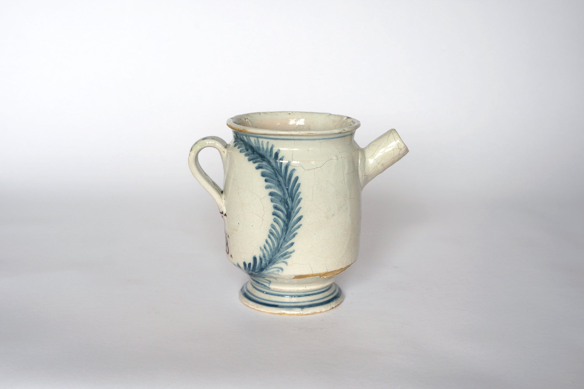 Euro Trash Vintage Hand-Painted Blue & Cream Ceramic Pitcher features an off-white body with blue leaf patterns, a side spout, handle, circular base, visible cracks, and a worn rim for antique appeal.