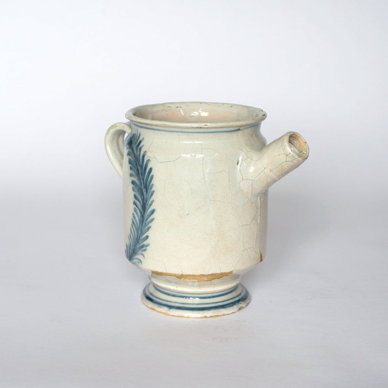 The Euro Trash Vintage Hand-Painted Blue & Cream Ceramic Pitcher features a cream base, blue leaf details, handle, short spout, and crackled glaze. It sits on a circular base against a plain white background.