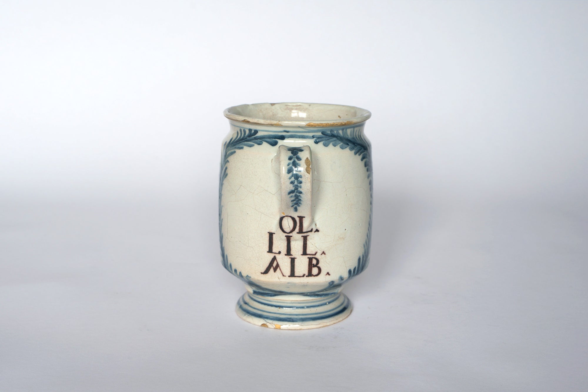 The Euro Trash Vintage Hand-Painted Blue & Cream Ceramic Pitcher features blue detailing, a pedestal base, and a weathered look with a cracked rim for added vintage charm. The inscription OL LIL ALB adorns its antique-inspired design.