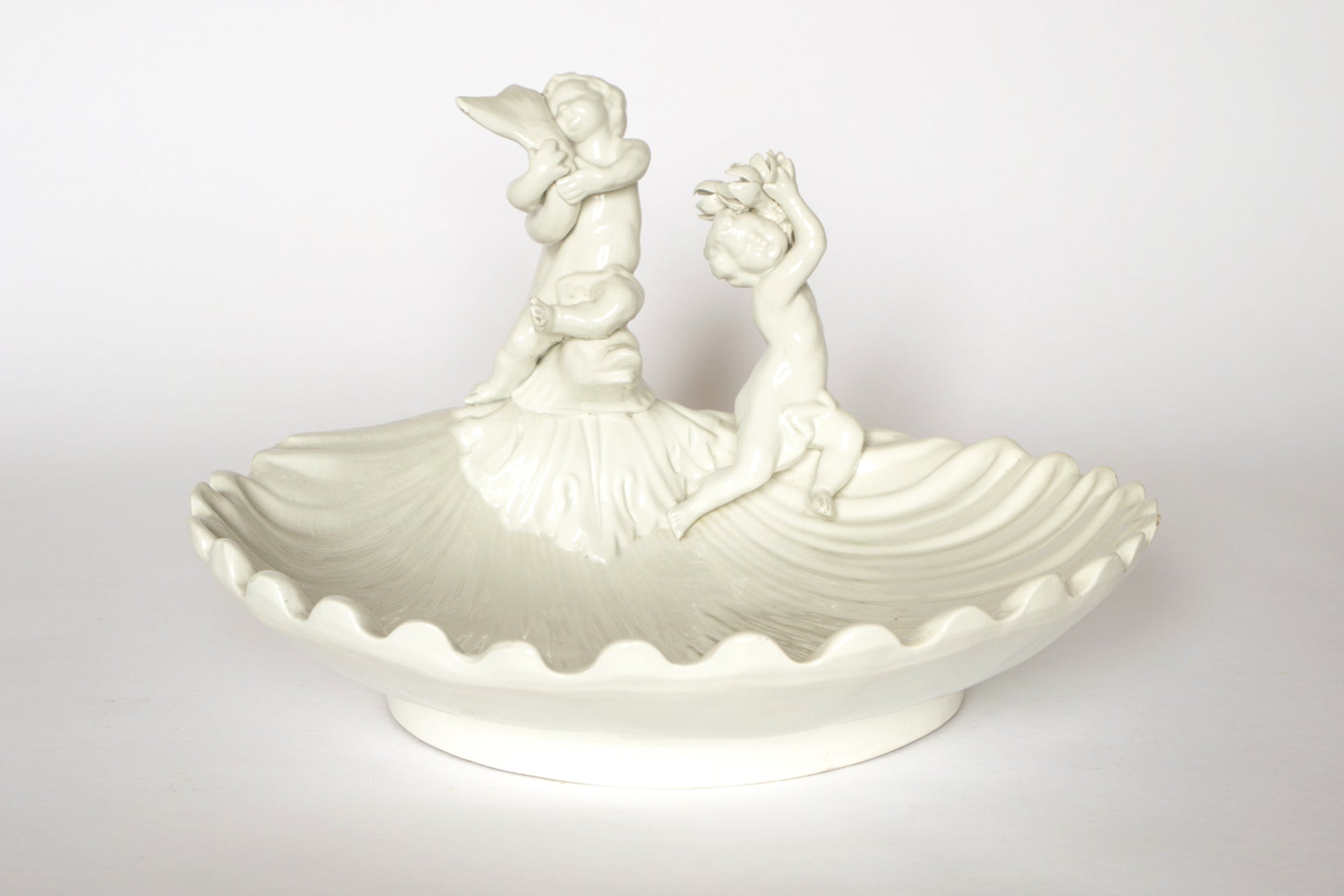 The Vintage White Ceramic Cherub Shell Dish by Euro Trash features a shell-shaped design with a child figurine and a seated cherub, making it an ideal centerpiece or charming trinket holder.
