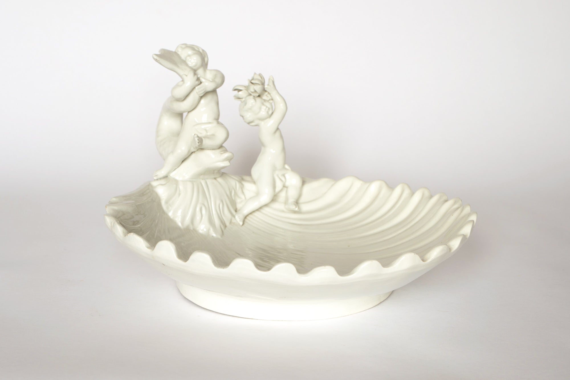 The Euro Trash Vintage White Ceramic Cherub Shell Dish features a wavy rim, glossy finish, and two cherubs—one sitting and playing a flute, the other standing and reaching. This charming dish is perfect as a decorative trinket holder.