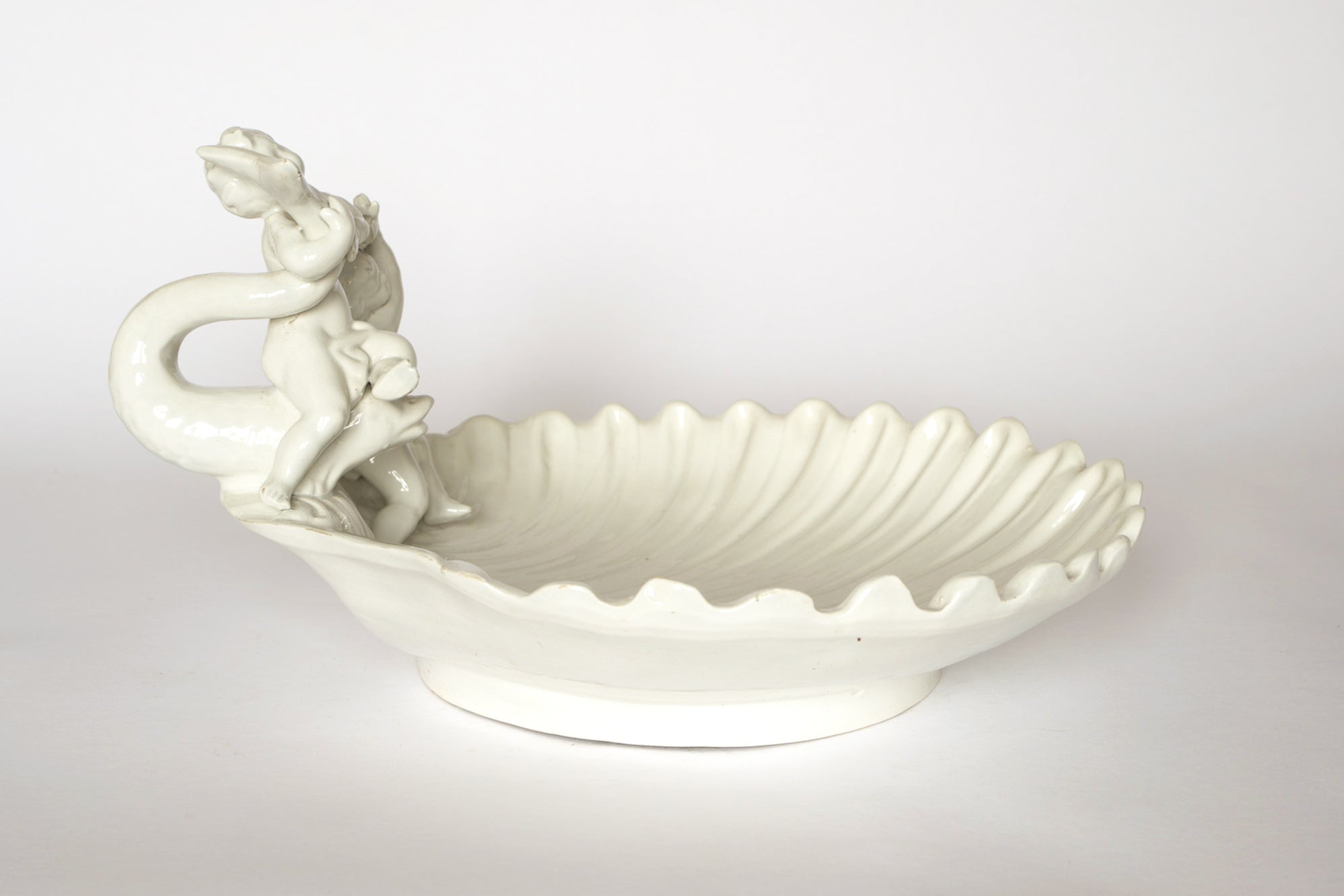 The Euro Trash Vintage White Ceramic Cherub Shell Dish features a glossy scallop shell shape with a sculpted cherub on a dolphin handle, making it an elegant decorative centerpiece.