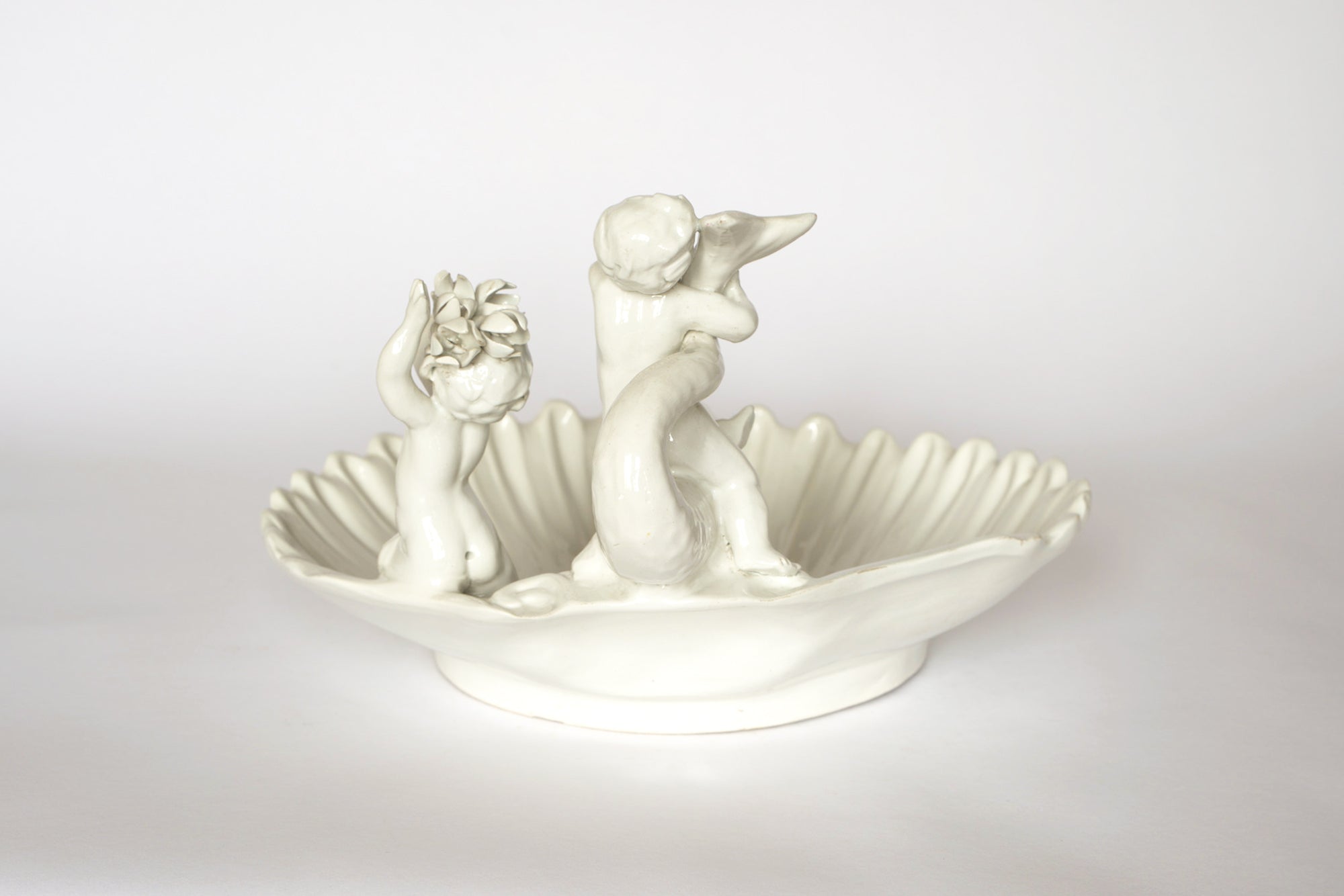 The Euro Trash Vintage White Ceramic Cherub Shell Dish features two tailed cherubs—one holding flowers, the other a horn—shaped as a scallop shell, making it a charming decorative centerpiece.