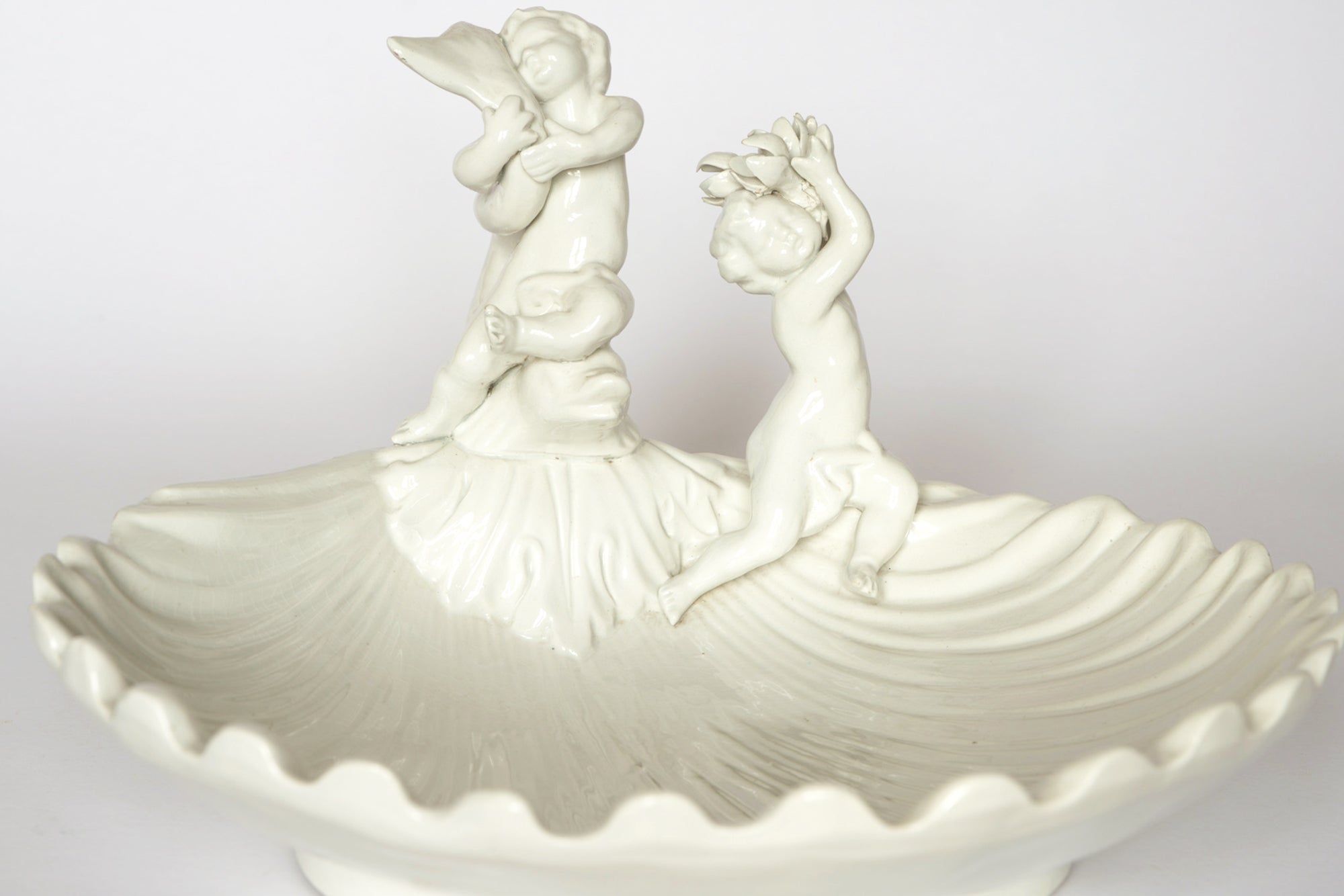 The Euro Trash Vintage White Ceramic Cherub Shell Dish features two cherubs—one standing and holding an object, the other reaching upward—beautifully integrated into its scalloped, shell-inspired design.