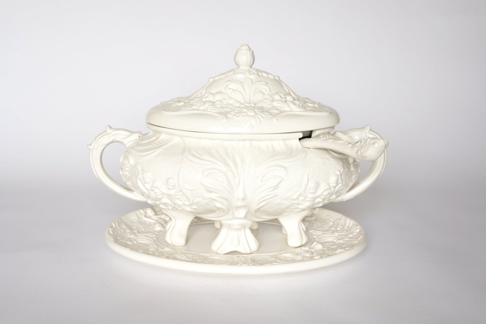 The Euro Trash Vintage White Ceramic Tureen with Underplate and Ladle features ornate floral and leaf designs, a matching lid with a raised handle, curved handles, and an included ladle—an elegant decorative soup serving set.