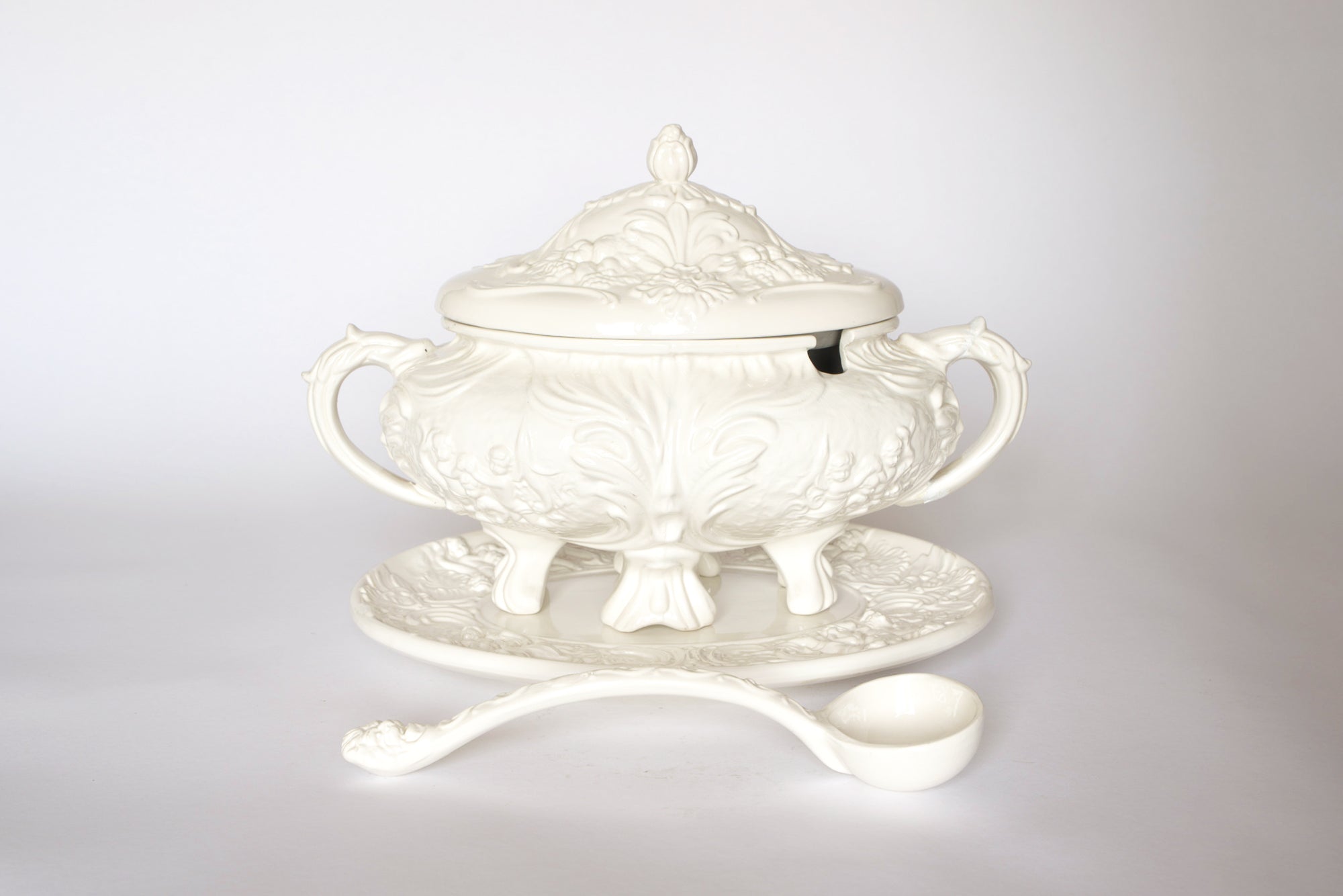 The Euro Trash Vintage White Ceramic Tureen with Underplate and Ladle features ornate handles, a matching lid and platter, and embossed patterns—perfect as a decorative soup serving set.