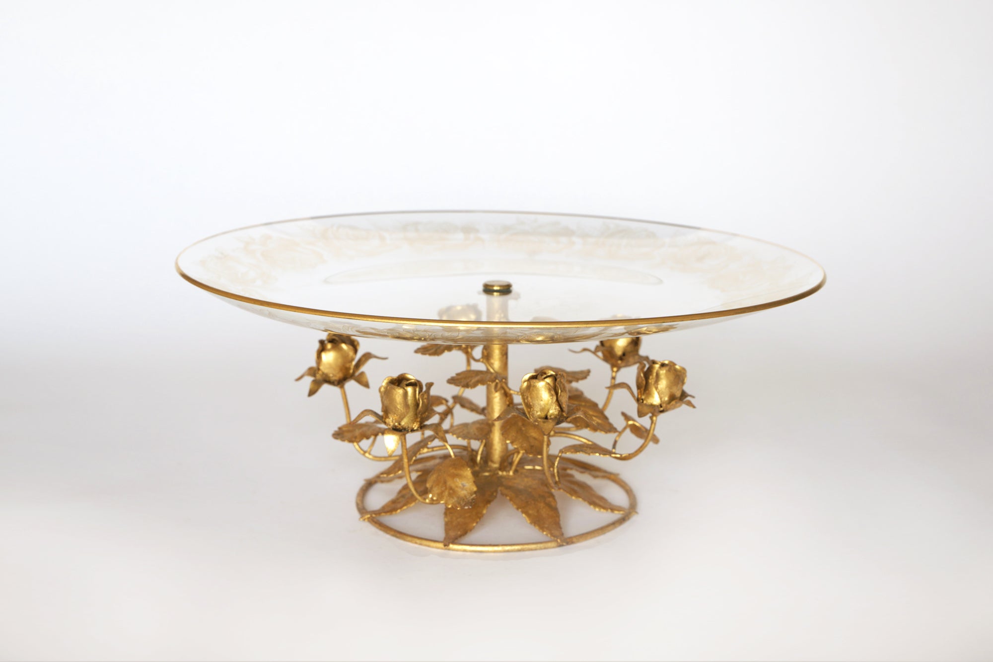 The Euro Trash Vintage Glass and Gilt Metal Rose Pedestal Cake Stand features a round clear glass top and an ornate gold-tone metal base with rose flowers and leaves, displayed against a plain white background.