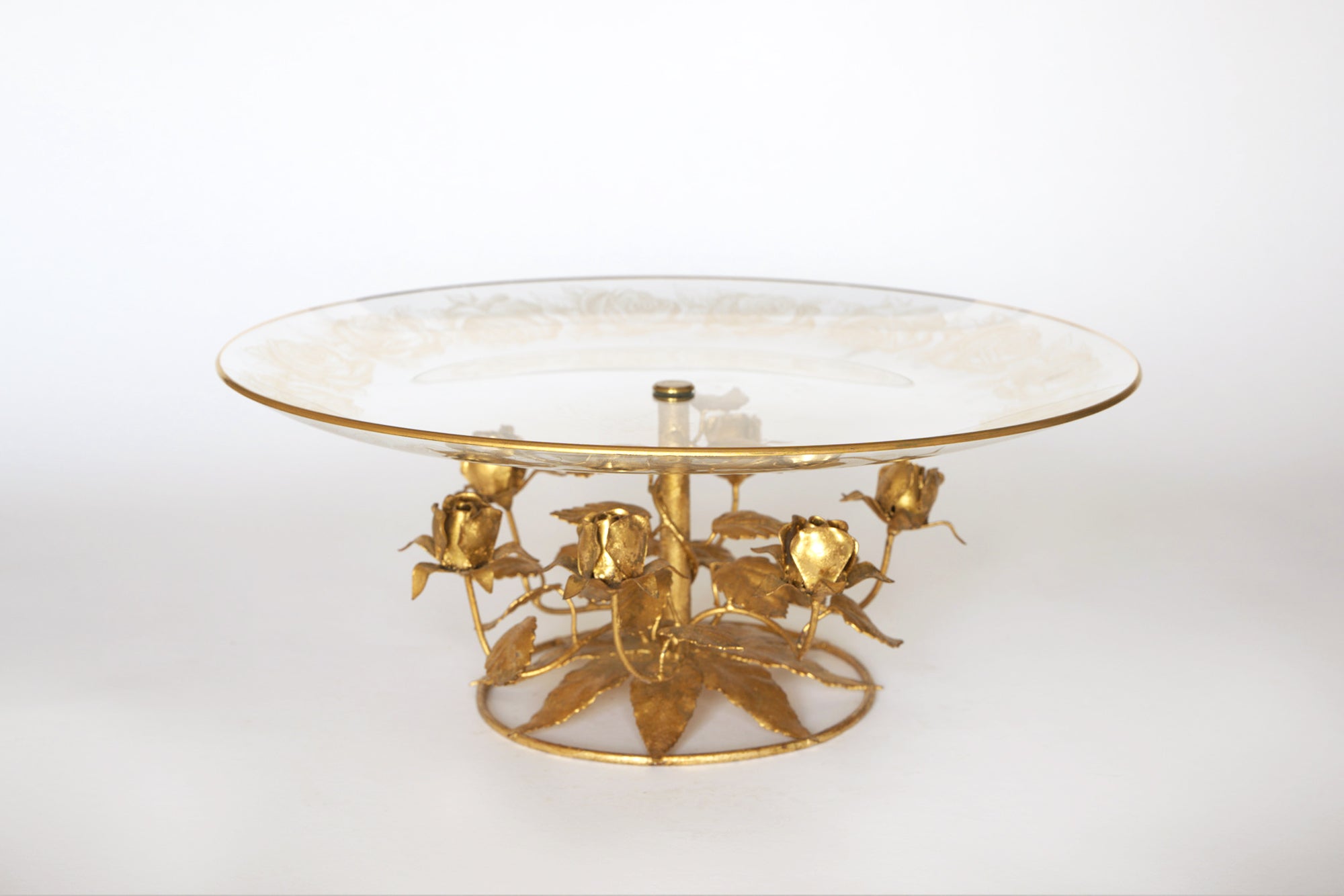 The Euro Trash Vintage Glass and Gilt Metal Rose Pedestal Cake Stand features a clear etched glass top and a gilt metal base with rose accents and leaf details, shown against a plain white background.