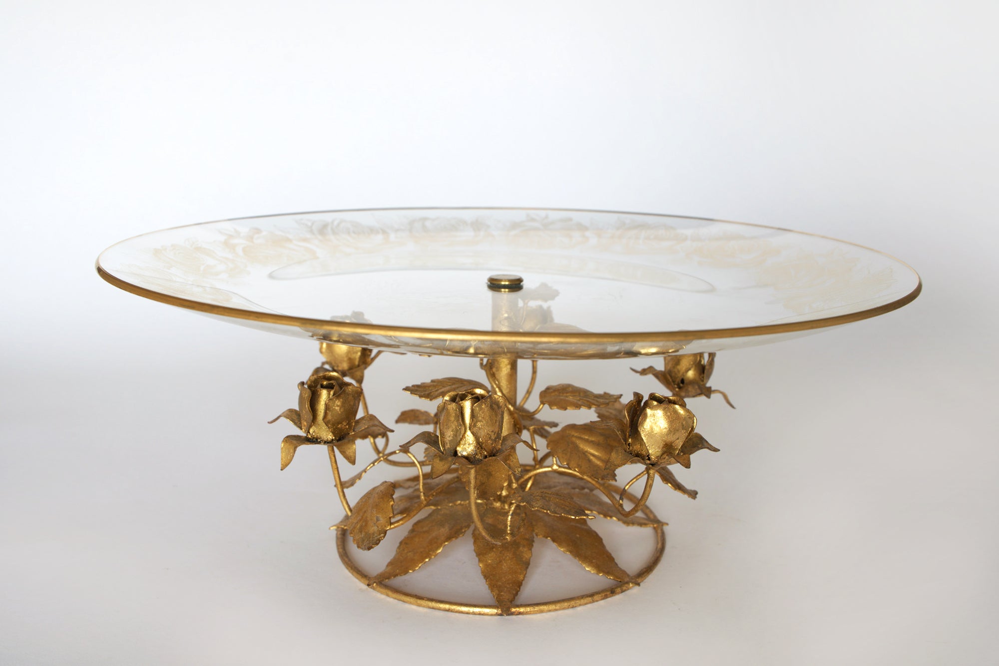 The Euro Trash Vintage Glass and Gilt Metal Rose Pedestal Cake Stand features a glass platter on a gilt metal base with sculpted roses and leaves, shown against a plain white background.