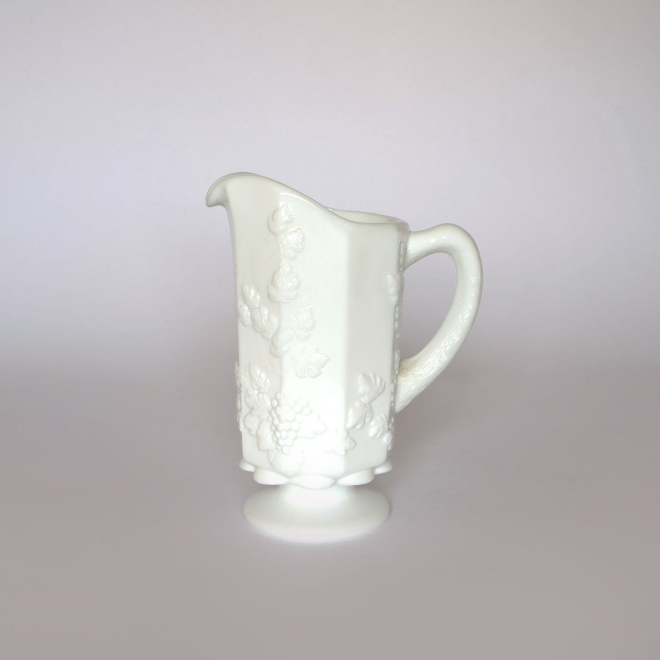 The Euro Trash Vintage White Milk Glass Pitcher features an embossed grapevine pattern, curved handle, and pedestal base—ideal for farmhouse decor and beautifully displayed against a light background.