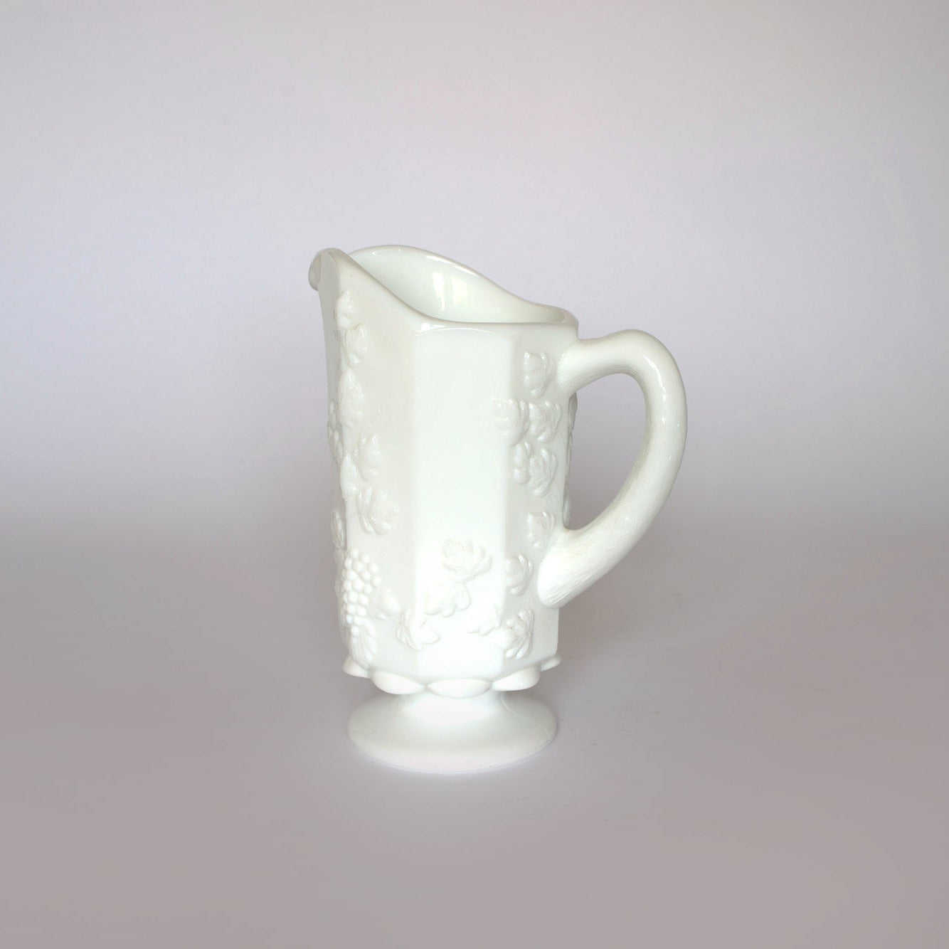 The Vintage White Milk Glass Pitcher by Euro Trash features embossed grapevine and floral patterns, a handle, and a pedestal base—an ideal farmhouse decor accent for any room. Shown against a plain light background.