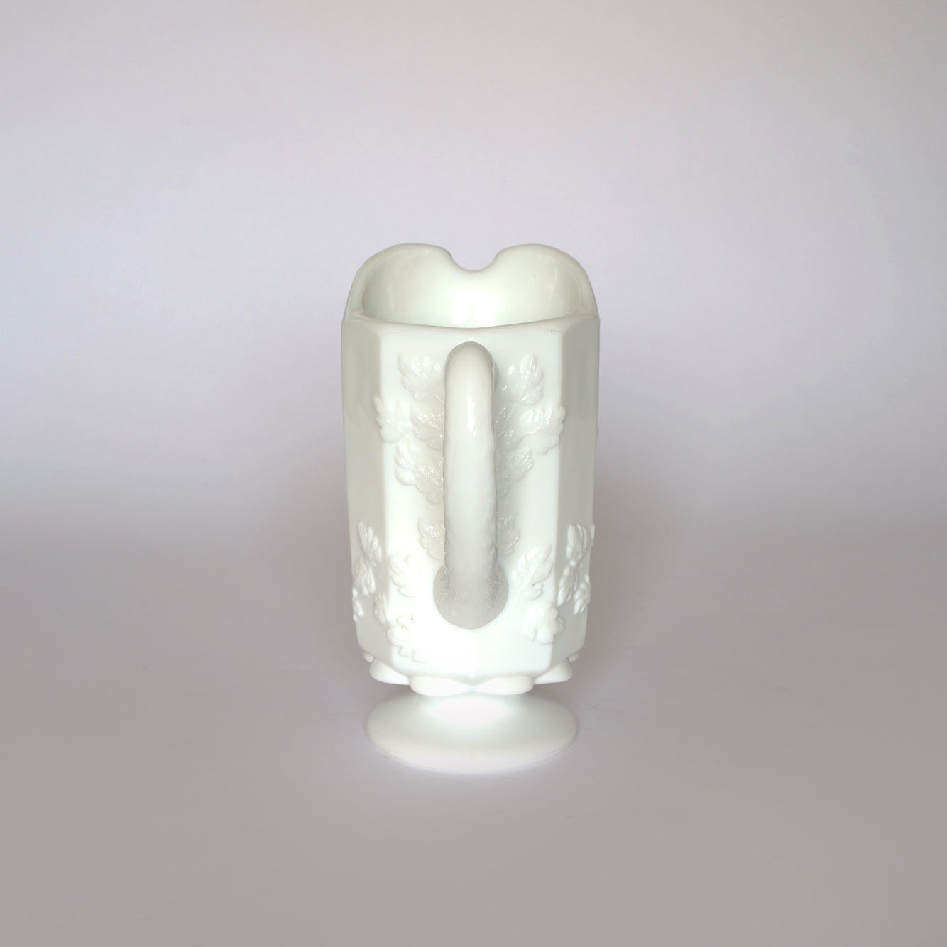 The Euro Trash Vintage White Milk Glass Pitcher features a classic embossed grapevine design, single handle, and round pedestal base—an ideal accent for farmhouse decor against any light gray backdrop.