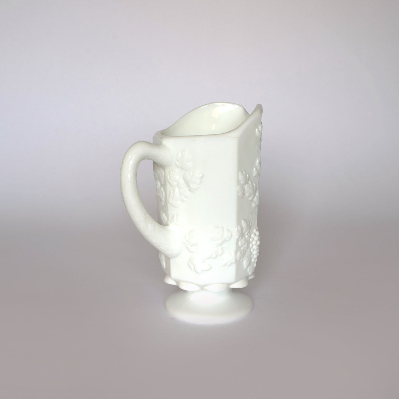 The Euro Trash Vintage White Milk Glass Pitcher features embossed grapevine patterns, a curved handle, and a pedestal base—ideal for farmhouse decor and photographed on a plain light gray background.