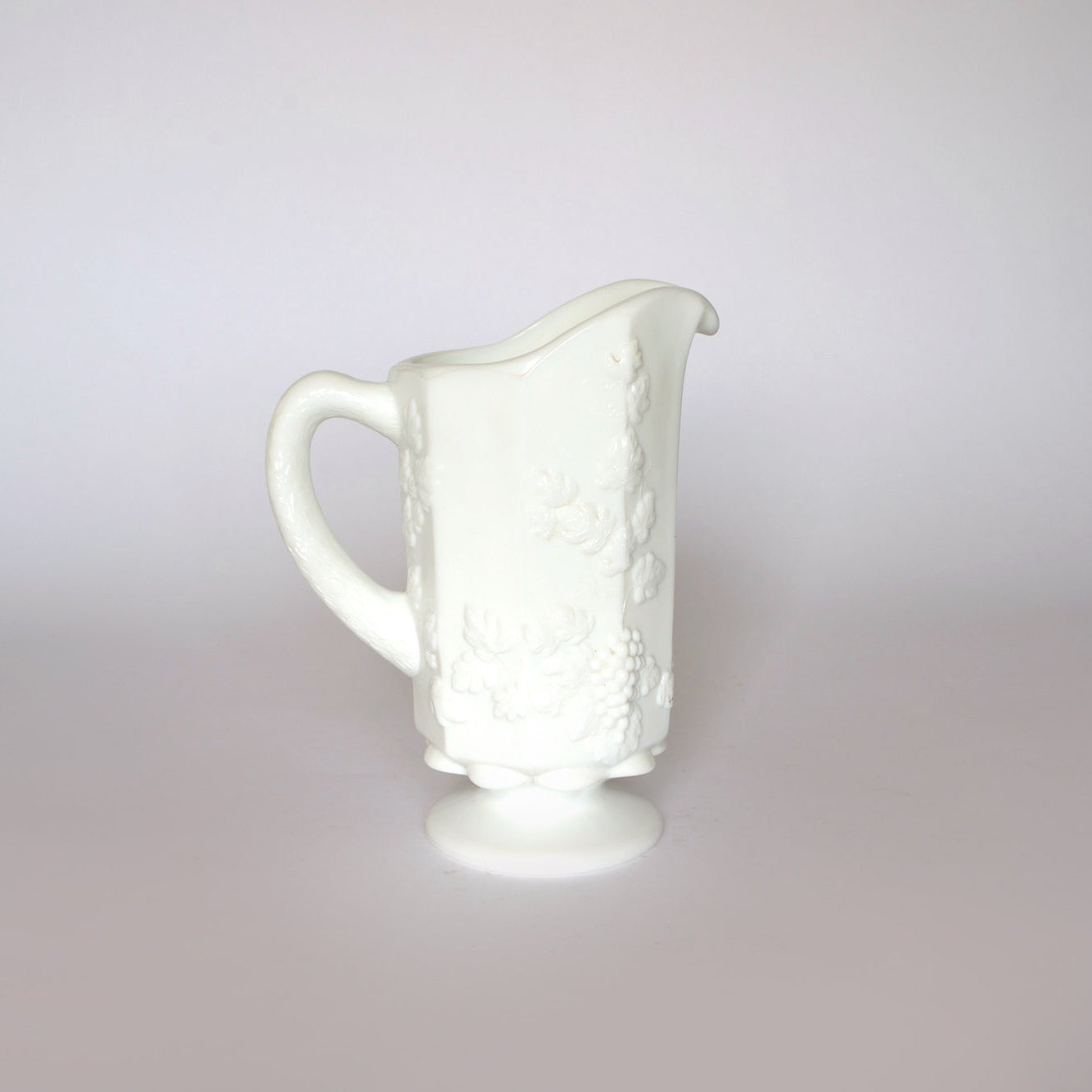 The Euro Trash Vintage White Milk Glass Pitcher features a single handle, embossed grapevine patterns, and a pedestal base—perfect for farmhouse decor—shown against a plain light gray background.