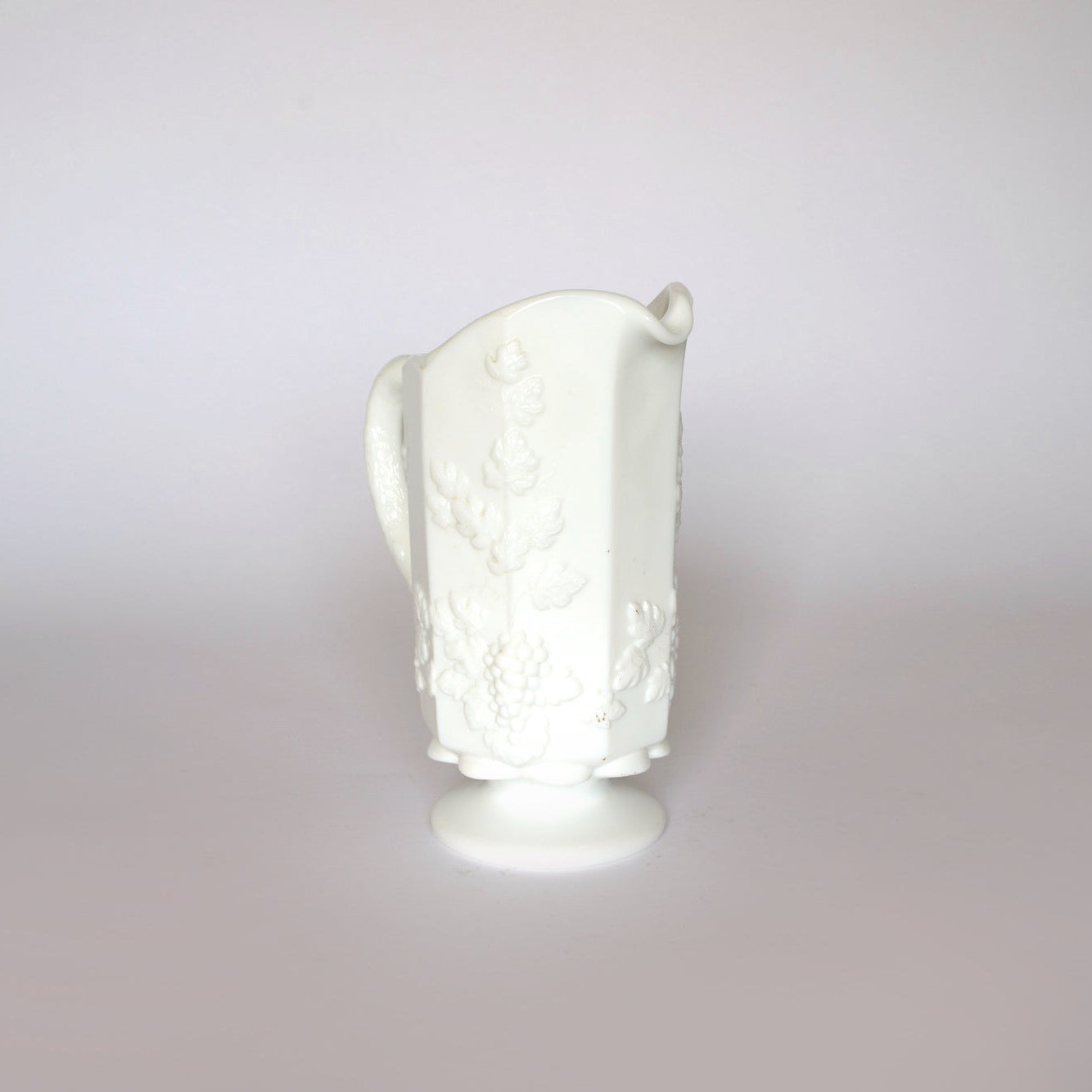 The Euro Trash Vintage White Milk Glass Pitcher features a curved handle, embossed grapevine design with leaves and grapes, and stands on a round pedestal base. Ideal for farmhouse decor, it’s shown against a plain light background.
