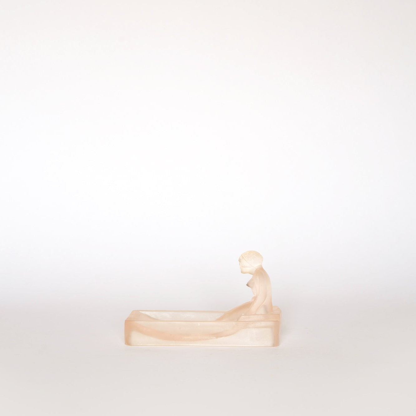 A small, pale frosted glass figure sits atop the edge of the rectangular Euro Trash Vintage Frosted Glass Figural Trinket Dish, set against a plain white background in soft, minimal lighting.