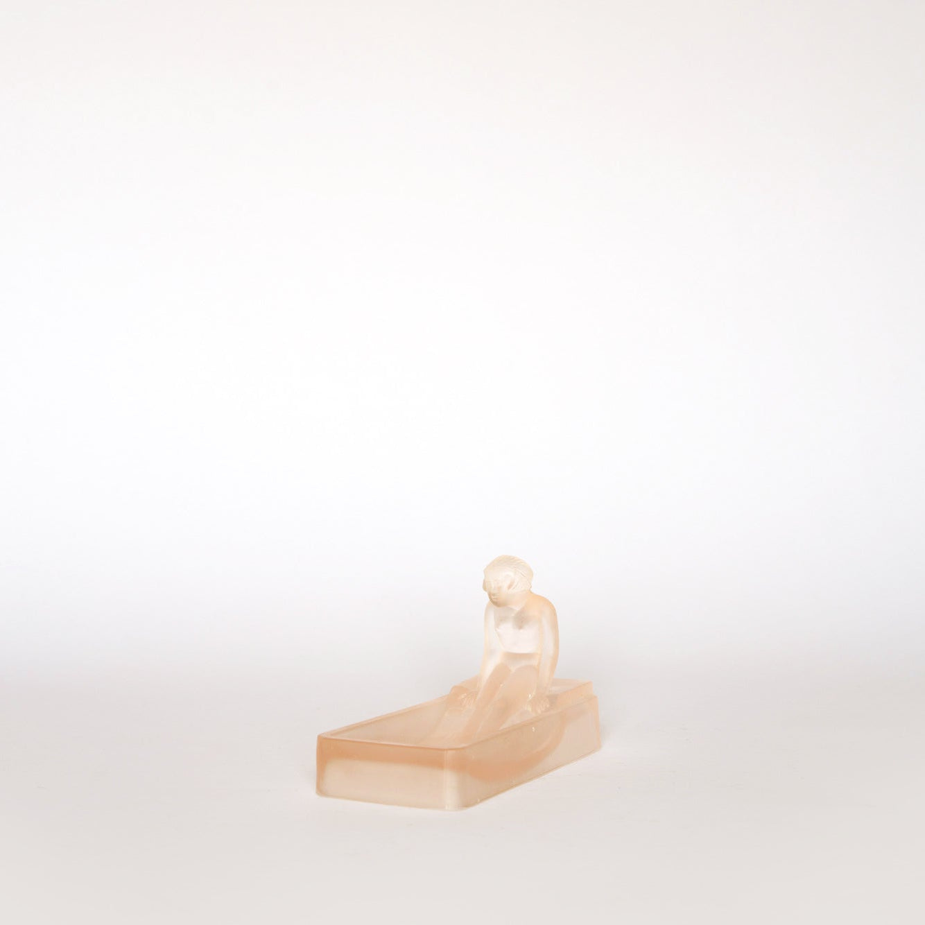 The Euro Trash Vintage Frosted Glass Figural Trinket Dish features a small, peach-toned seated figure in Art Deco style on a rectangular base, displayed against a plain white background.