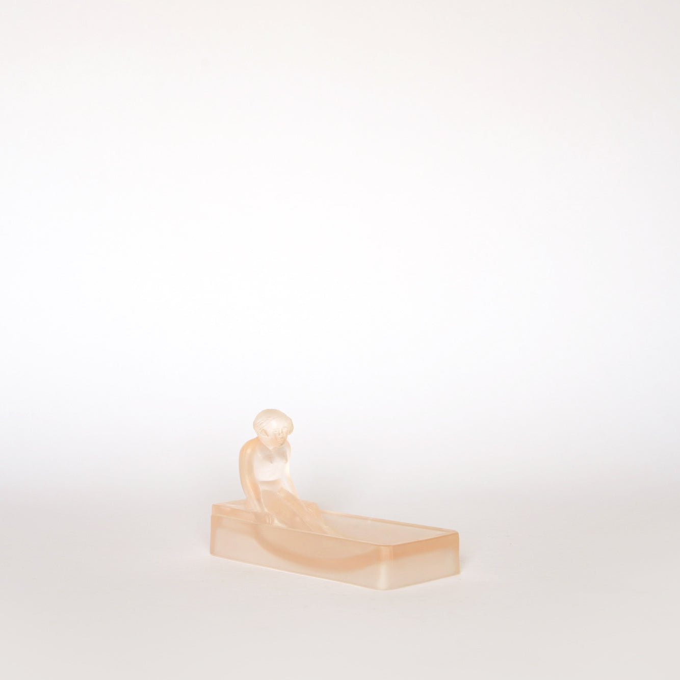 The Euro Trash Vintage Frosted Glass Figural Trinket Dish showcases a peach-hued, translucent seated figure on a rectangular block in Art Deco style, highlighted against a plain white background.