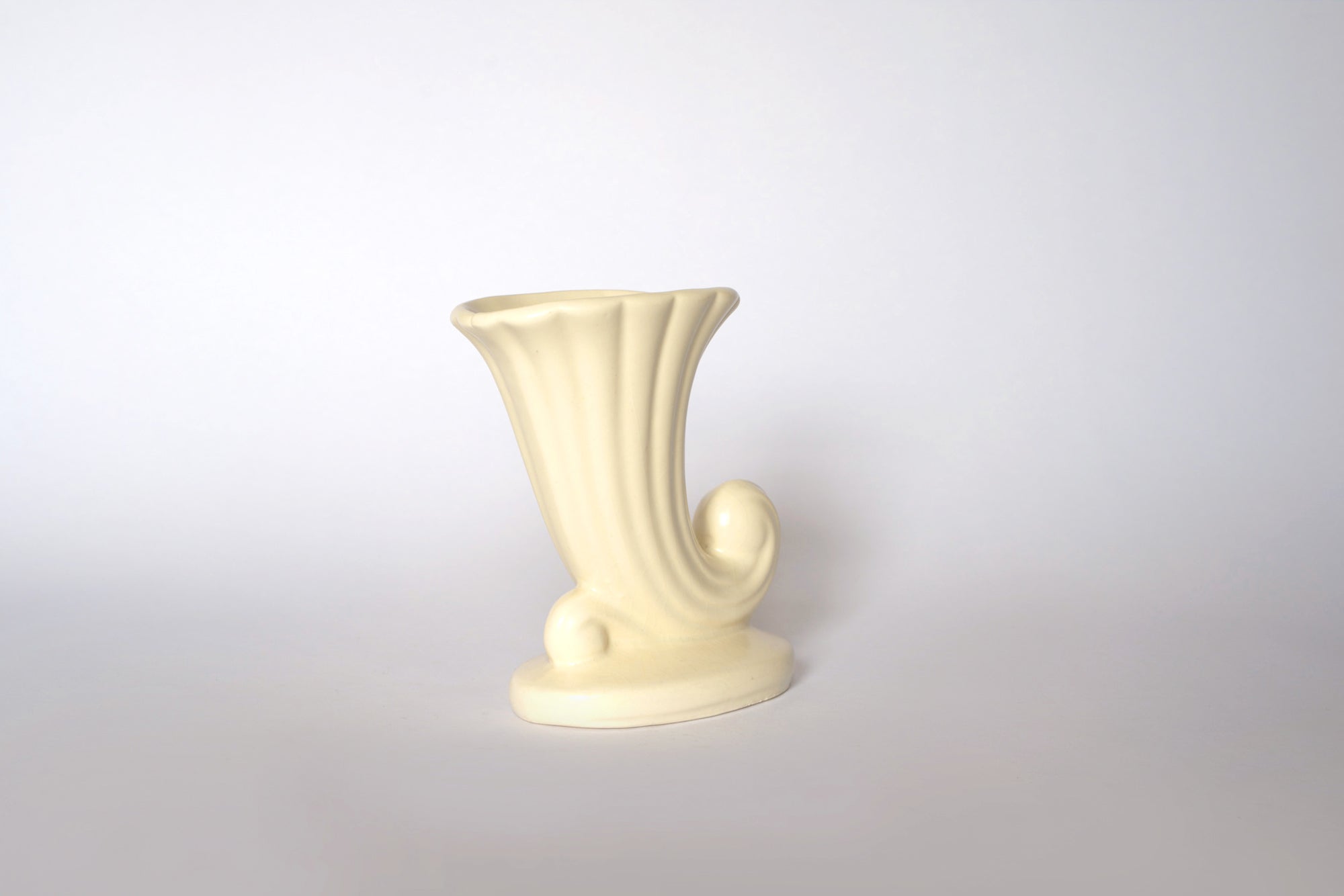 The Euro Trash Vintage Cream Ceramic Cornucopia Vase features a fluted opening and curved decorative base, perfect for cottage-style décor, shown against a plain white background.