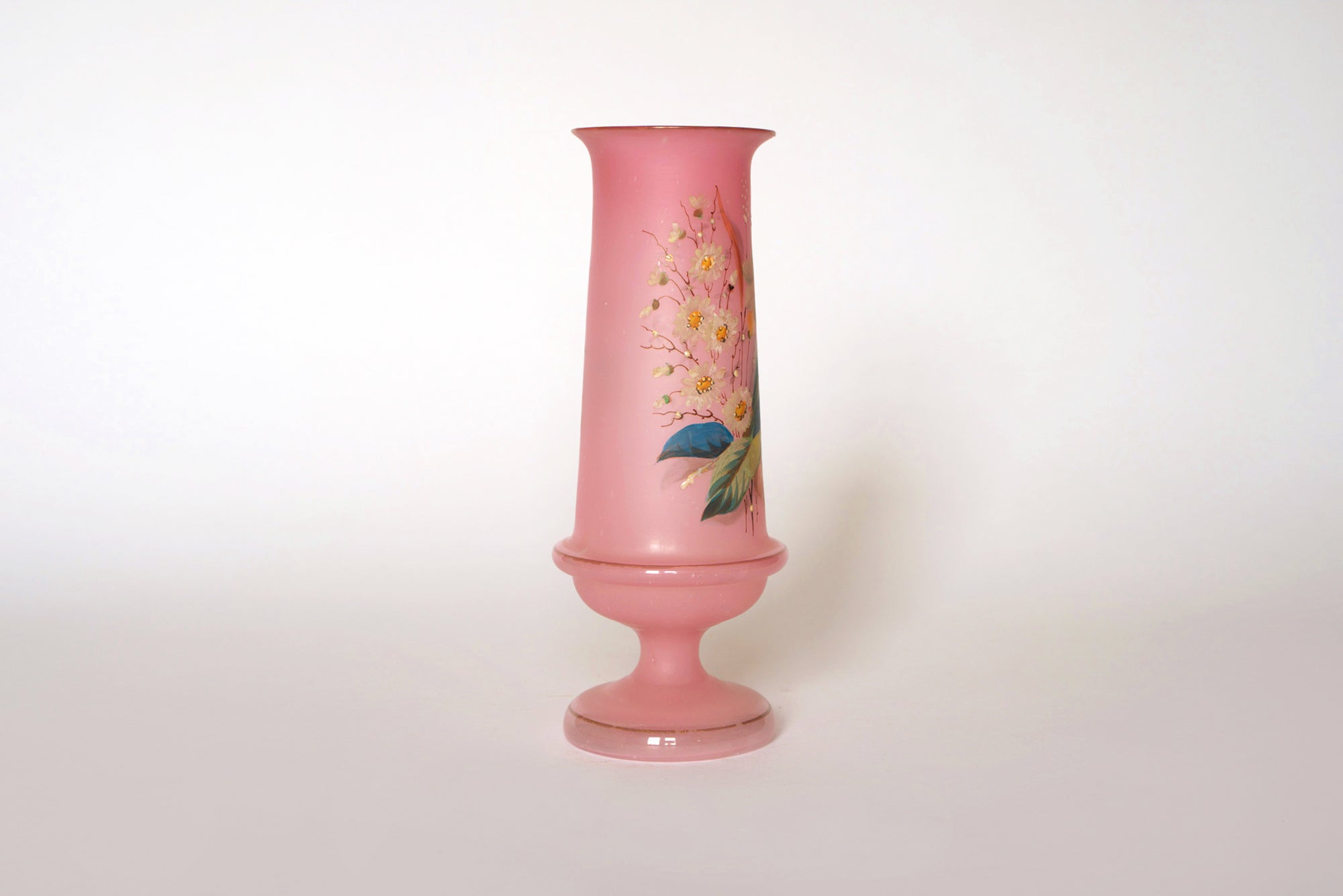 The Euro Trash Vintage Pink Floral Glass Vase features a tall, wide-based pink design with a narrow neck and hand-painted yellow and pink flowers with green leaves on a plain white background.