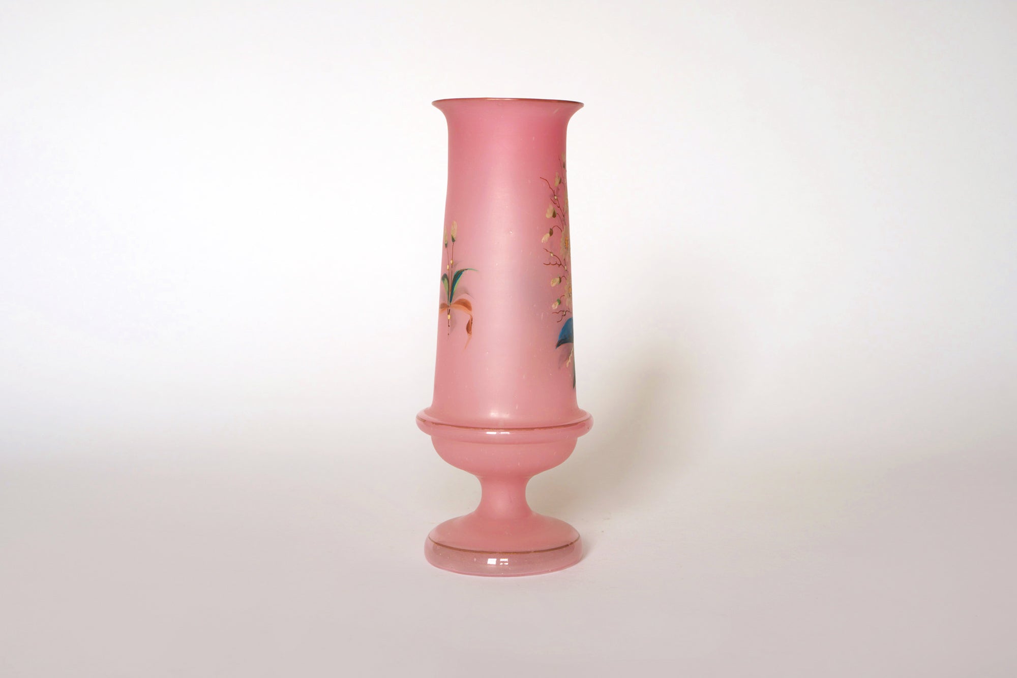 The Euro Trash Vintage Pink Floral Glass Vase features a tall, narrow neck and wide base, adorned with delicate hand-painted flowers and leaves, set against a plain white background.
