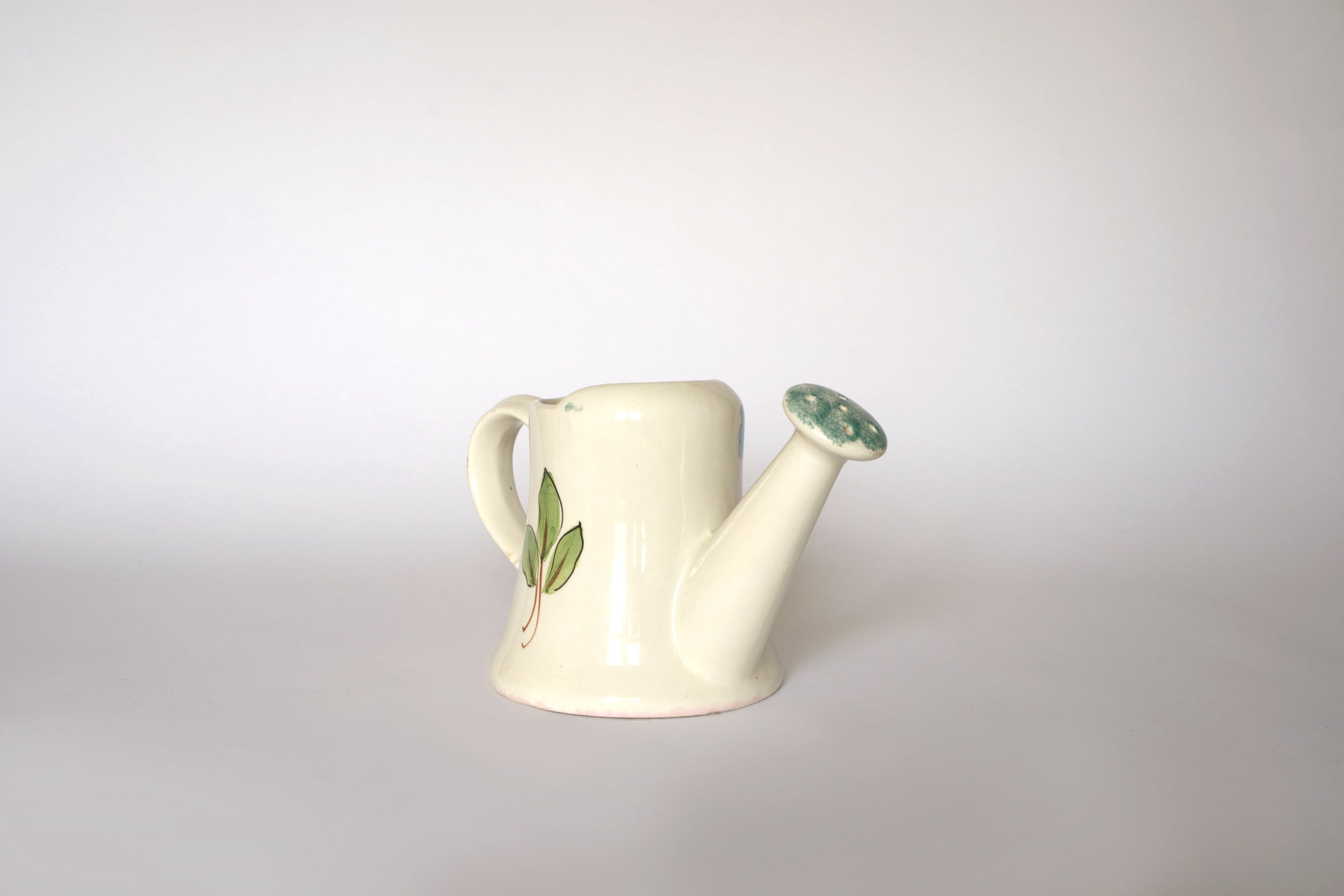 The Euro Trash Vintage Hand-Painted Floral Ceramic Watering Can is a small, cream-colored can with green leaf accents and a short spout, featuring a floral design on a plain white background.