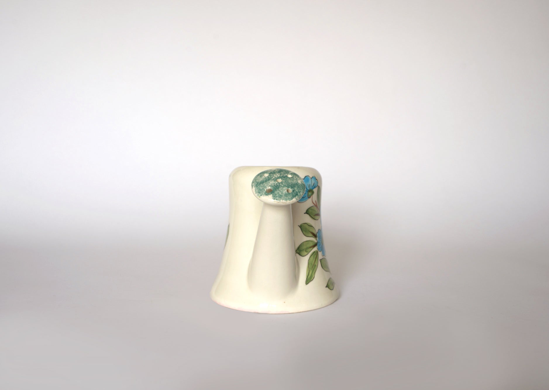 The Euro Trash Vintage Hand-Painted Floral Ceramic Watering Can, cream-colored with a blue flower and green leaves, and a flared base, is shown upside down on a plain white background.