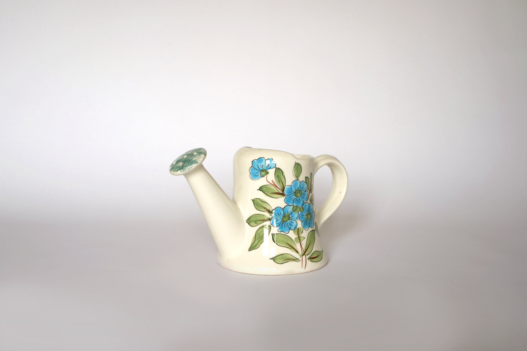 The Euro Trash Vintage Hand-Painted Floral Ceramic Watering Can, cream-colored with blue flowers and green leaves, has a handle and spout and makes a charming decorative accent against a plain white background.