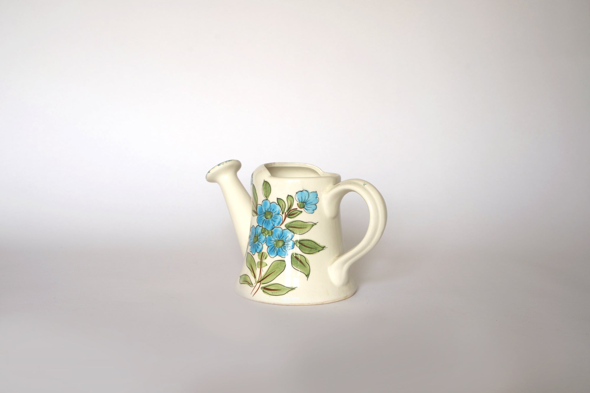 The Euro Trash Vintage Hand-Painted Floral Ceramic Watering Can, adorned with blue flowers and green leaves, features a handle and spout, making it a charming decor accent against a light background.