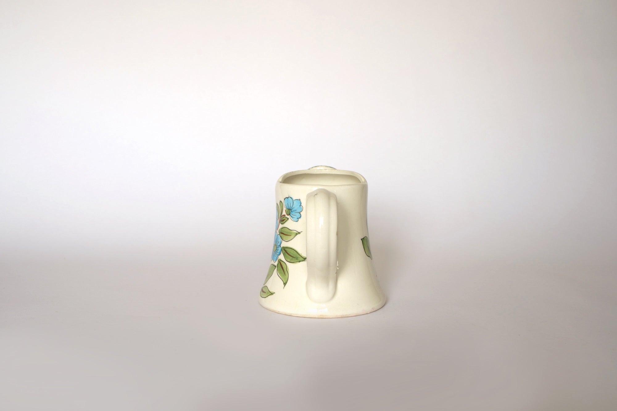 The Euro Trash Vintage Hand-Painted Floral Ceramic Watering Can features blue flowers and green leaves, shown from the back with its handle and lid visible against a plain light background.