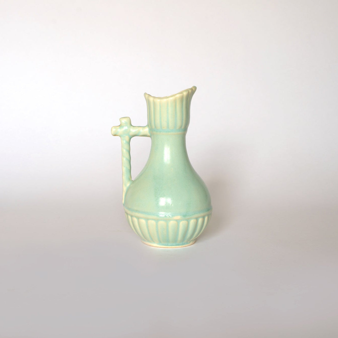 The Euro Trash Vintage Glazed Ceramic Pitcher Vase features a light green ribbed body, elongated neck, and ornate handle—an ideal decorative accent for any space, displayed on a plain white background.