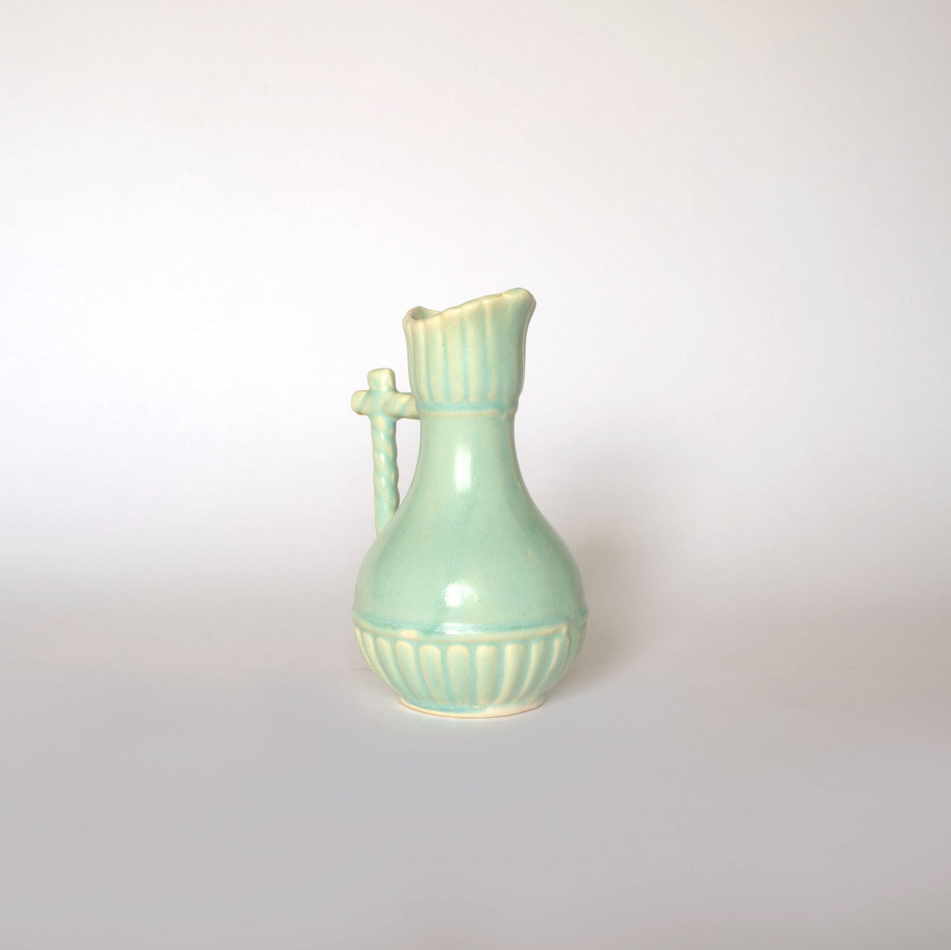 The Euro Trash Vintage Glazed Ceramic Pitcher Vase features a light green finish, fluted neck, rounded body, decorative handle, and ribbed detailing—a charming accent vase for any space. Shown on a plain white background.