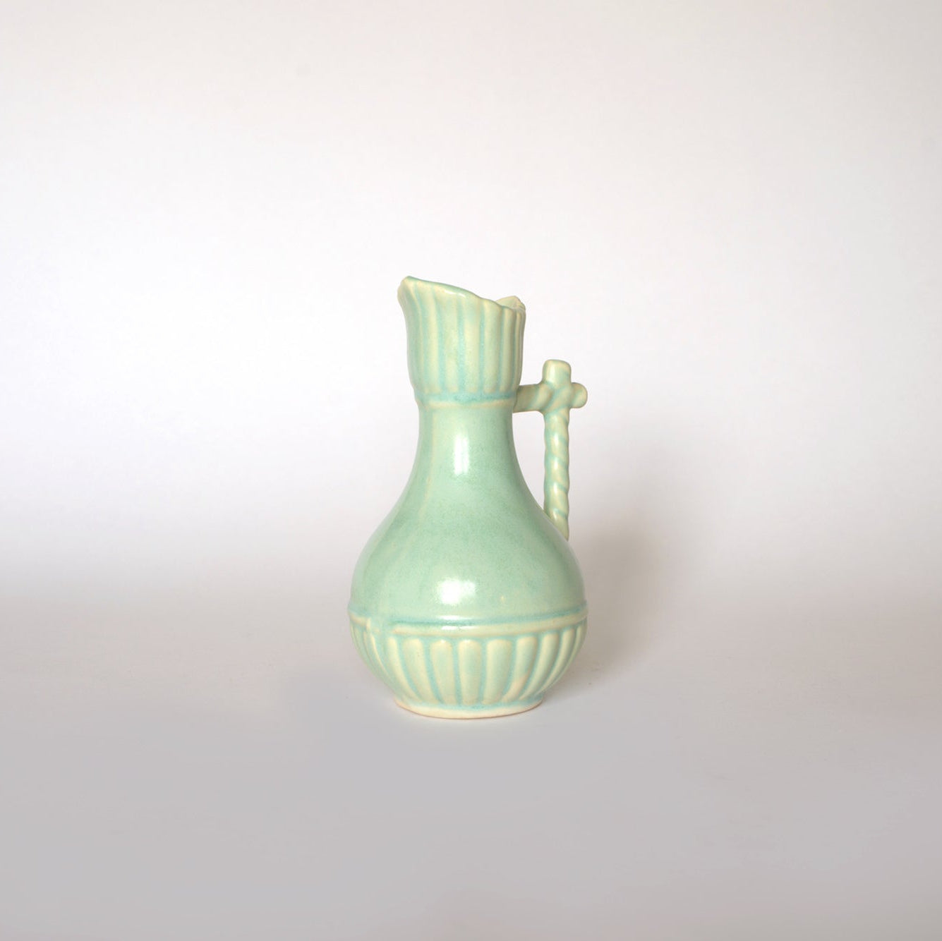 The Vintage Glazed Ceramic Pitcher Vase by Euro Trash features a light green finish, curved body, vertical ridges near the top and bottom, and a twisted decorative handle—perfect as a charming accent on any plain white background.