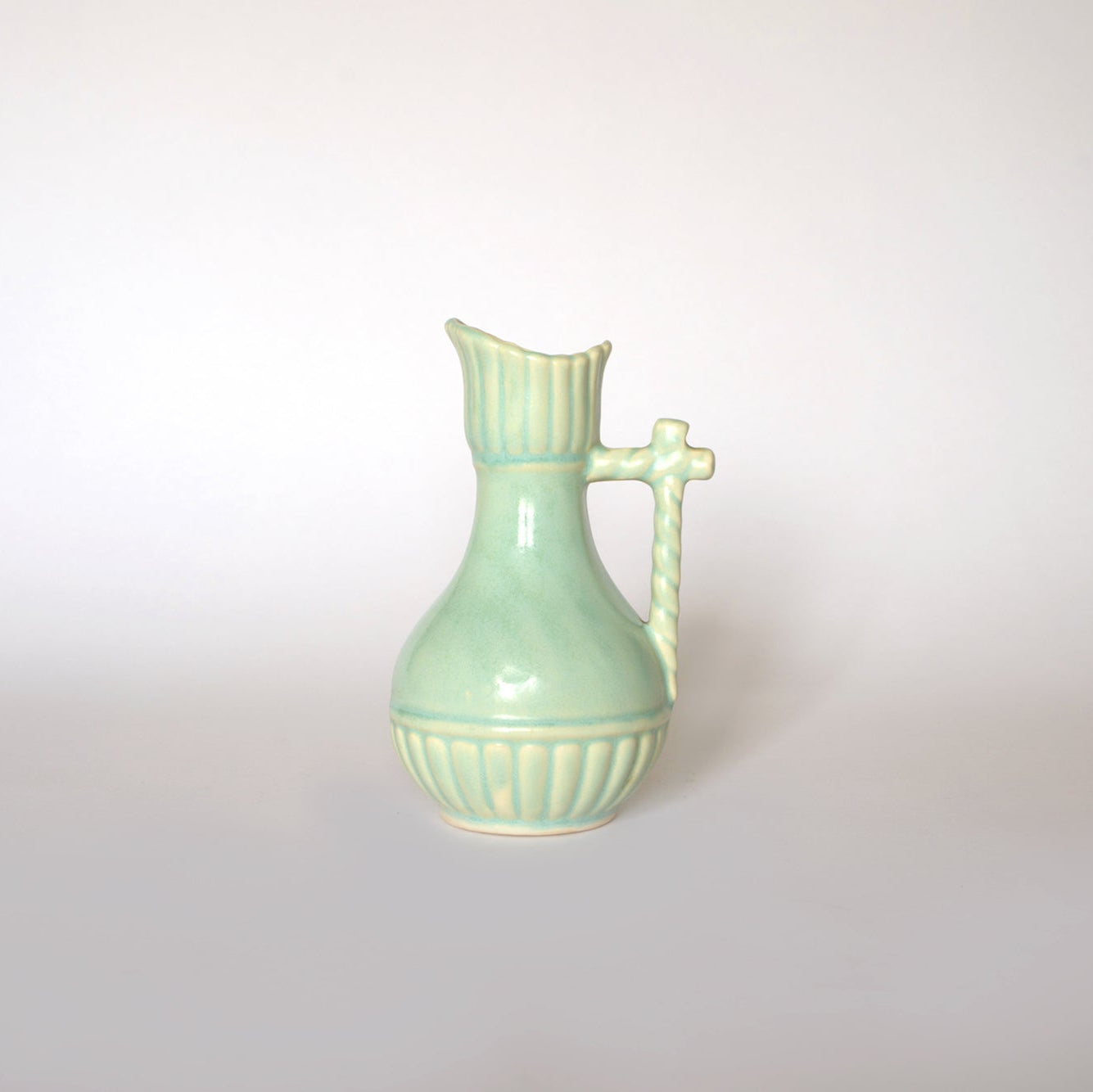 The Euro Trash Vintage Glazed Ceramic Pitcher Vase features a ribbed pale green ceramic body, narrow neck, flared spout, and twisted handle, making it a charming decorative accent against any plain light background.
