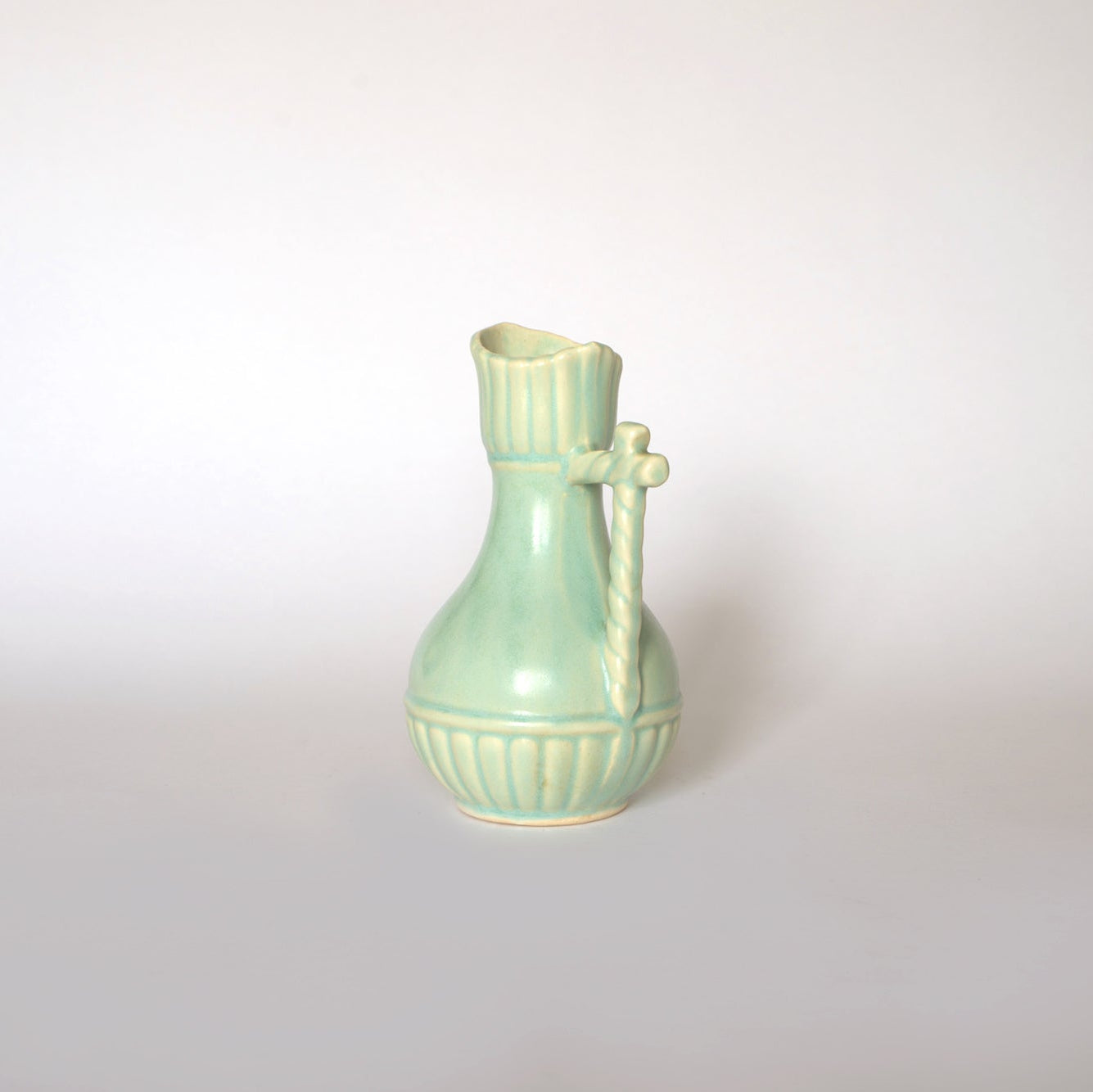 The Euro Trash Vintage Glazed Ceramic Pitcher Vase, featuring a pale green glaze, round base, narrow neck, wavy rim, and twisted side handle, makes a charming decorative accent against a plain light background.