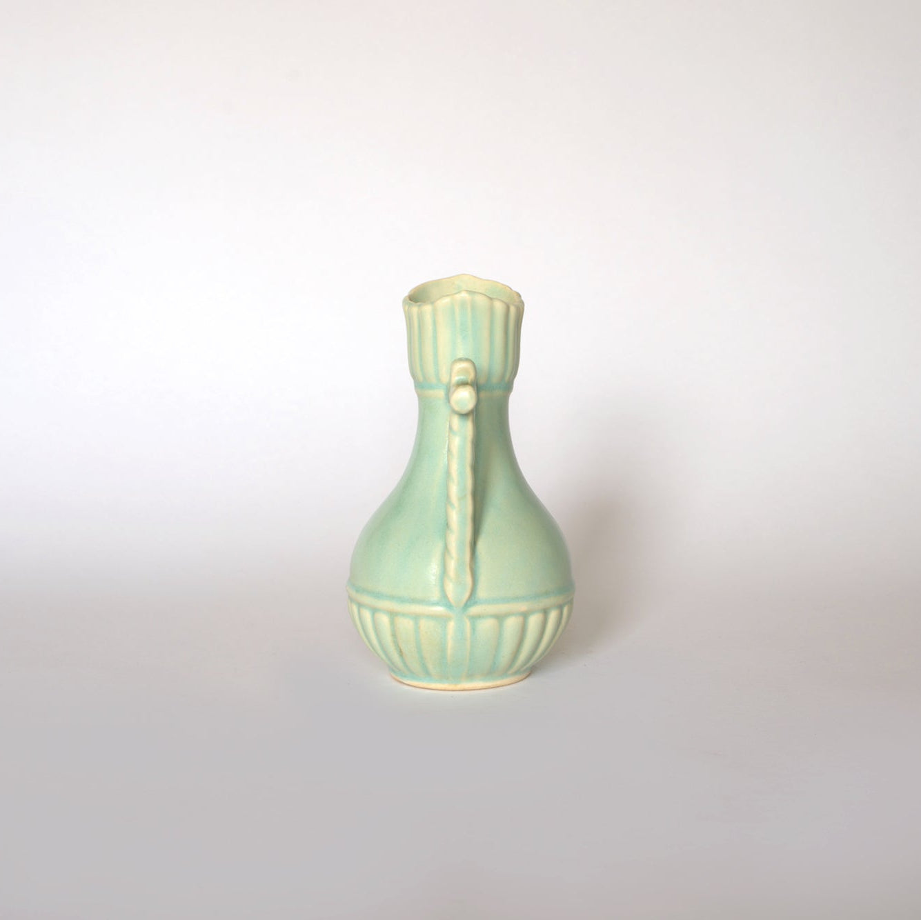The Euro Trash Vintage Glazed Ceramic Pitcher Vase features pale mint green ceramic, a rounded base, narrow neck, and decorative vertical ridges with rope-like details, making it a stylish accent piece against a plain white background.