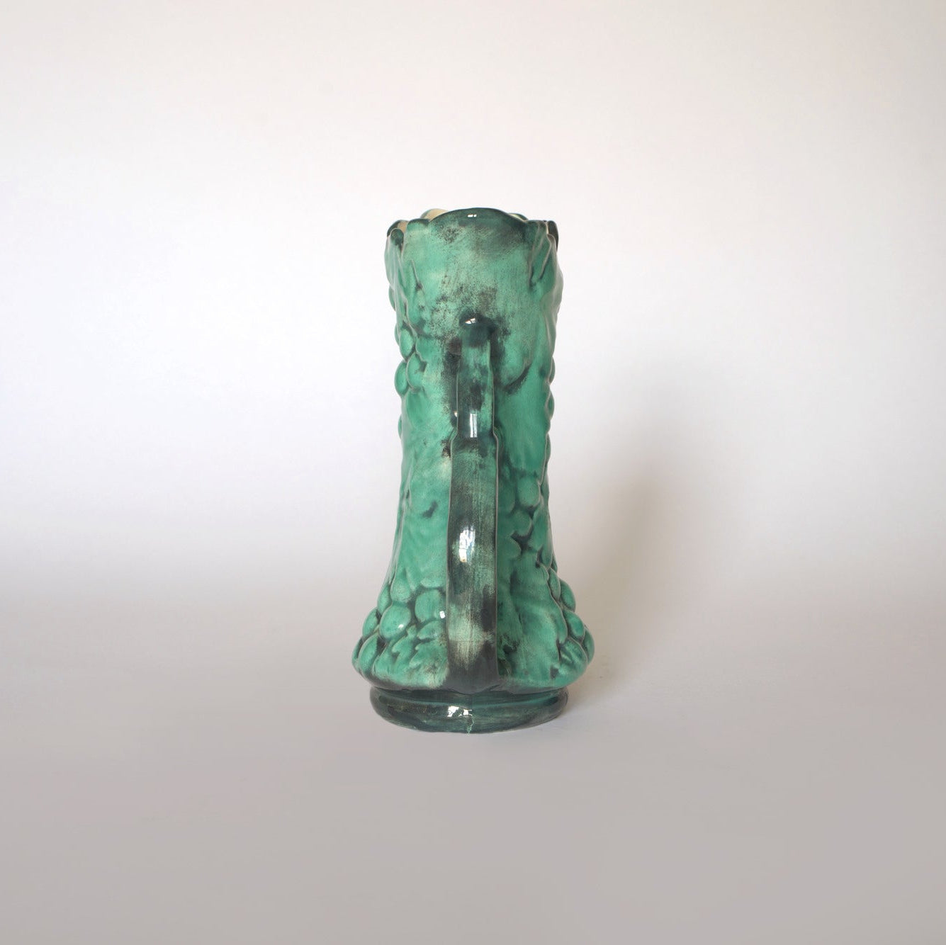 The Euro Trash Vintage Green Majolica Style Grape Motif Ceramic Pitcher, with handle and raised grapes and leaves design, is shown from the back against a plain light background in this photo.