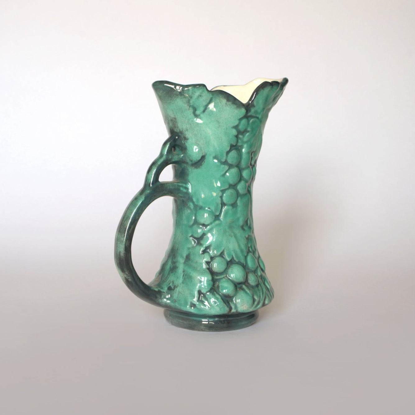 The Euro Trash Vintage Green Majolica Style Grape Motif Ceramic Pitcher, featuring a textured surface, wavy rim, and thick curved handle, stands against a plain light background, highlighting its charming organic design.