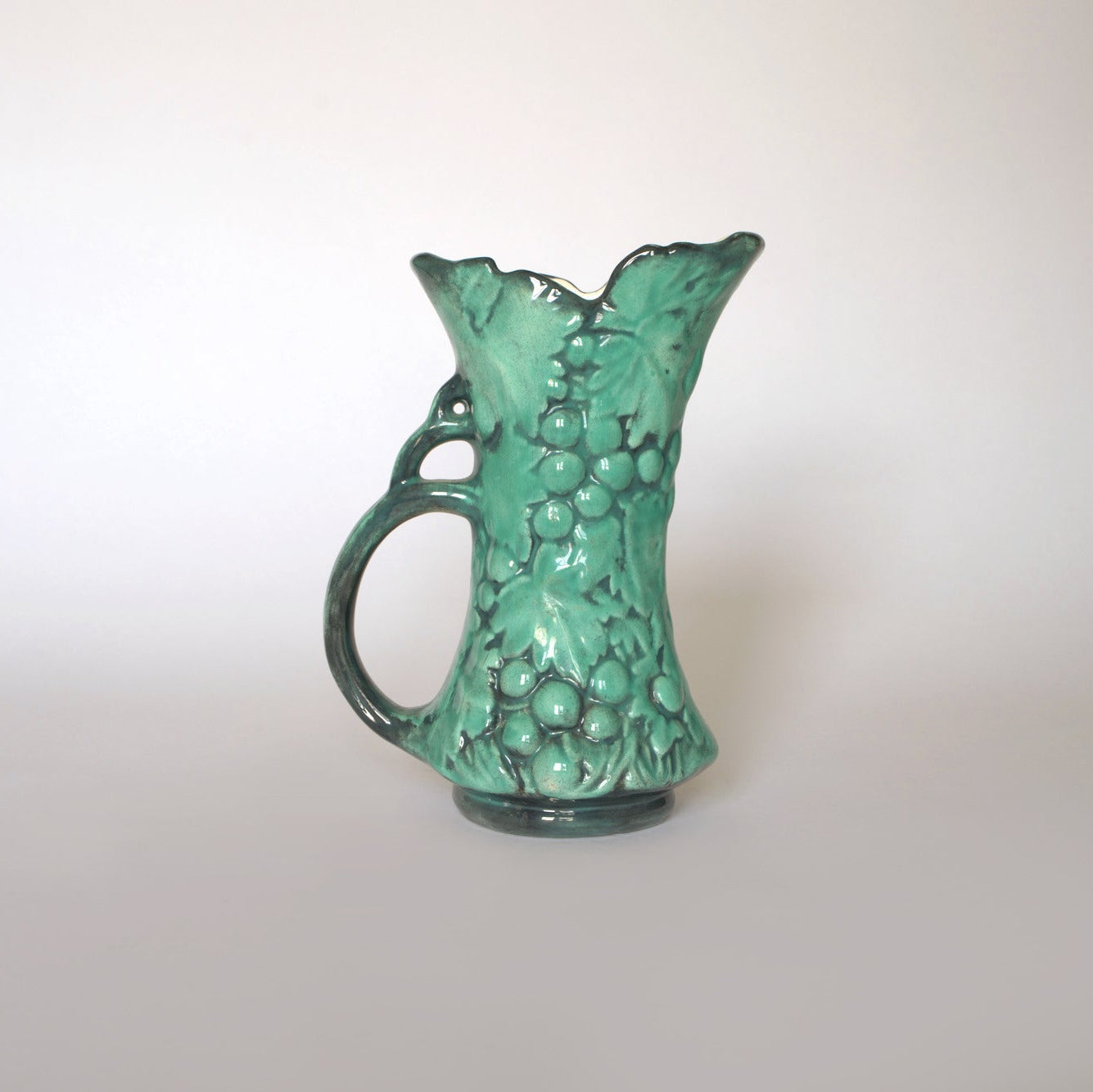 The Euro Trash Vintage Green Majolica Style Grape Motif Ceramic Pitcher features an ornate handle and raised grapes and leaves, beautifully detailed against its glossy green finish and shown on a plain white background.