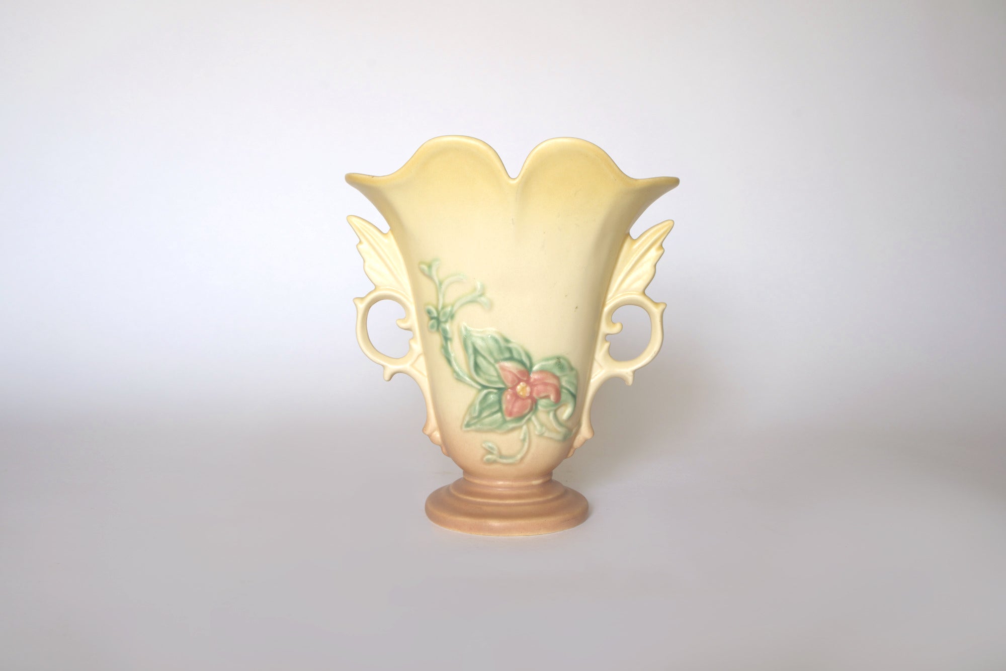 The Vintage Hull Floral Vase by Euro Trash features ornate handles, a scalloped rim, and a floral design with green leaves and a pink flower on a beige-yellow gradient background, evoking classic Hull Pottery.