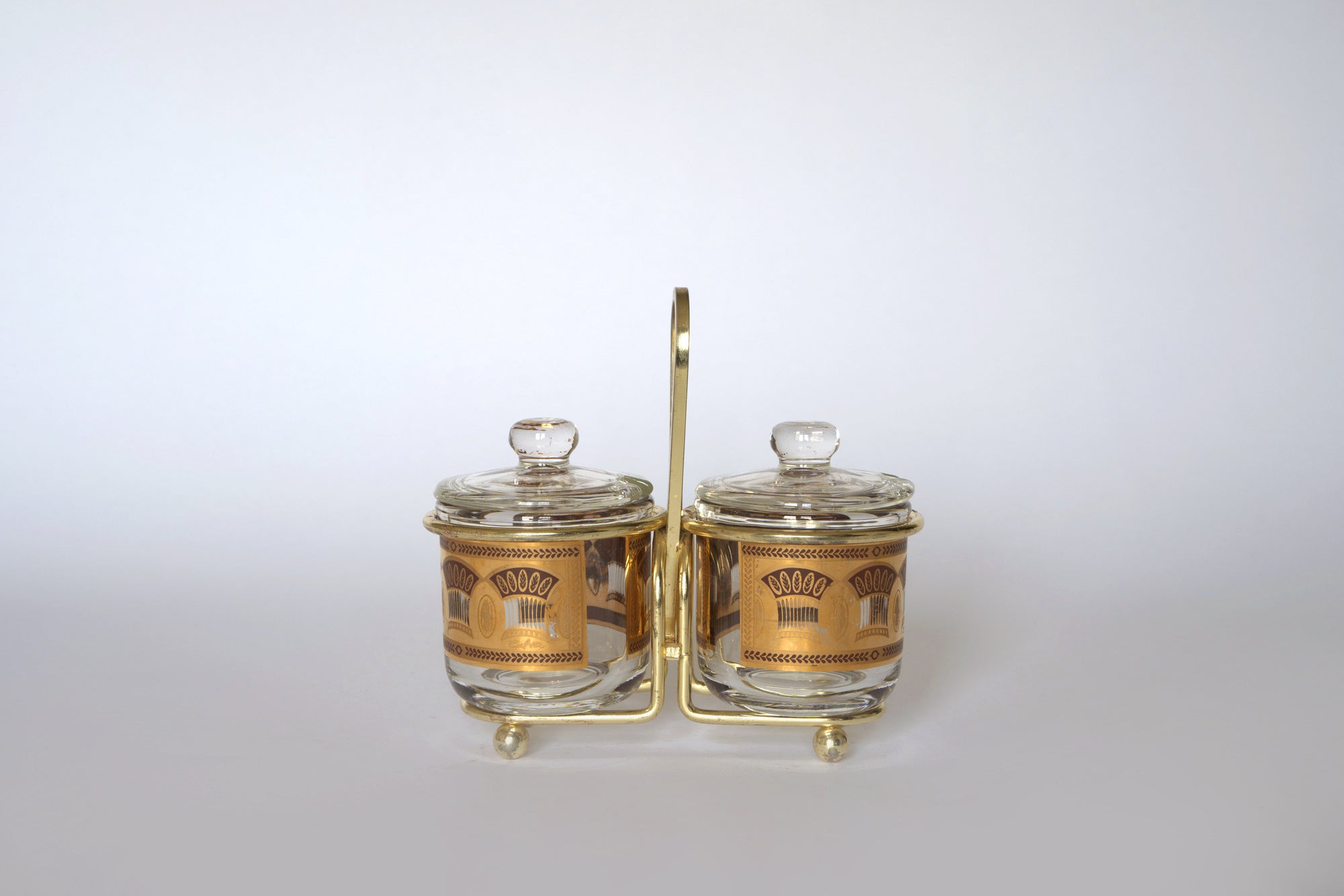 The Euro Trash Vintage Gold Caddy Double Glass Jar Set features two jars with gold patterns, clear lids, and a gold caddy—perfect for mid-century kitchen decor—shown on a plain white background.